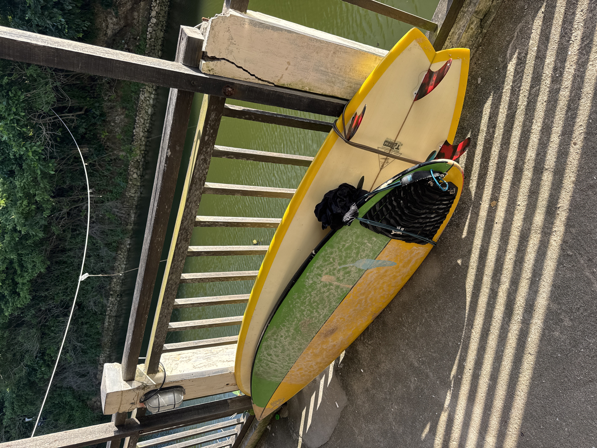 The image displays an outdoor scene featuring two surfboards propped against a wooden railing, overlooking a calm body of green water surrounded by lush vegetation. The setting appears to be a bridge or an elevated walkway, likely in a tropical location such as Kuta Selatan, Indonesia.
Two surfboards are the primary subjects. The top board is predominantly yellow with a cream-colored deck and features two striking red and black patterned fins near its tail. The word "TEAM" is faintly visible on its deck. Beneath it, a second surfboard is partially seen, sporting a green and yellow striped design, lightly dusted with sand or dirt. A black bag, possibly containing wetsuits or gear, and a black surfboard leash are attached to the boards.
The surfboards lean against a weathered wooden railing, constructed from sturdy posts and horizontal slats. The railing itself shows signs of age and exposure to the elements. A concrete structural element of the bridge, positioned next to the railing, exhibits a noticeable crack. A small, caged light fixture is attached to one of the wooden posts. A thin white cable or wire traverses the left side of the frame, extending towards the distant bank.
Below the railing, a tranquil body of green water flows. Its banks are rugged, lined with rocks and dense green trees and shrubs, characteristic of a natural waterway. The time of day is indicated by strong, elongated shadows cast by the wooden railing onto the concrete ground, suggesting either early morning or late afternoon sunlight. The weather is bright and clear. No people are visible, and the scene is static, implying the surfboards are temporarily stored rather than actively being used.