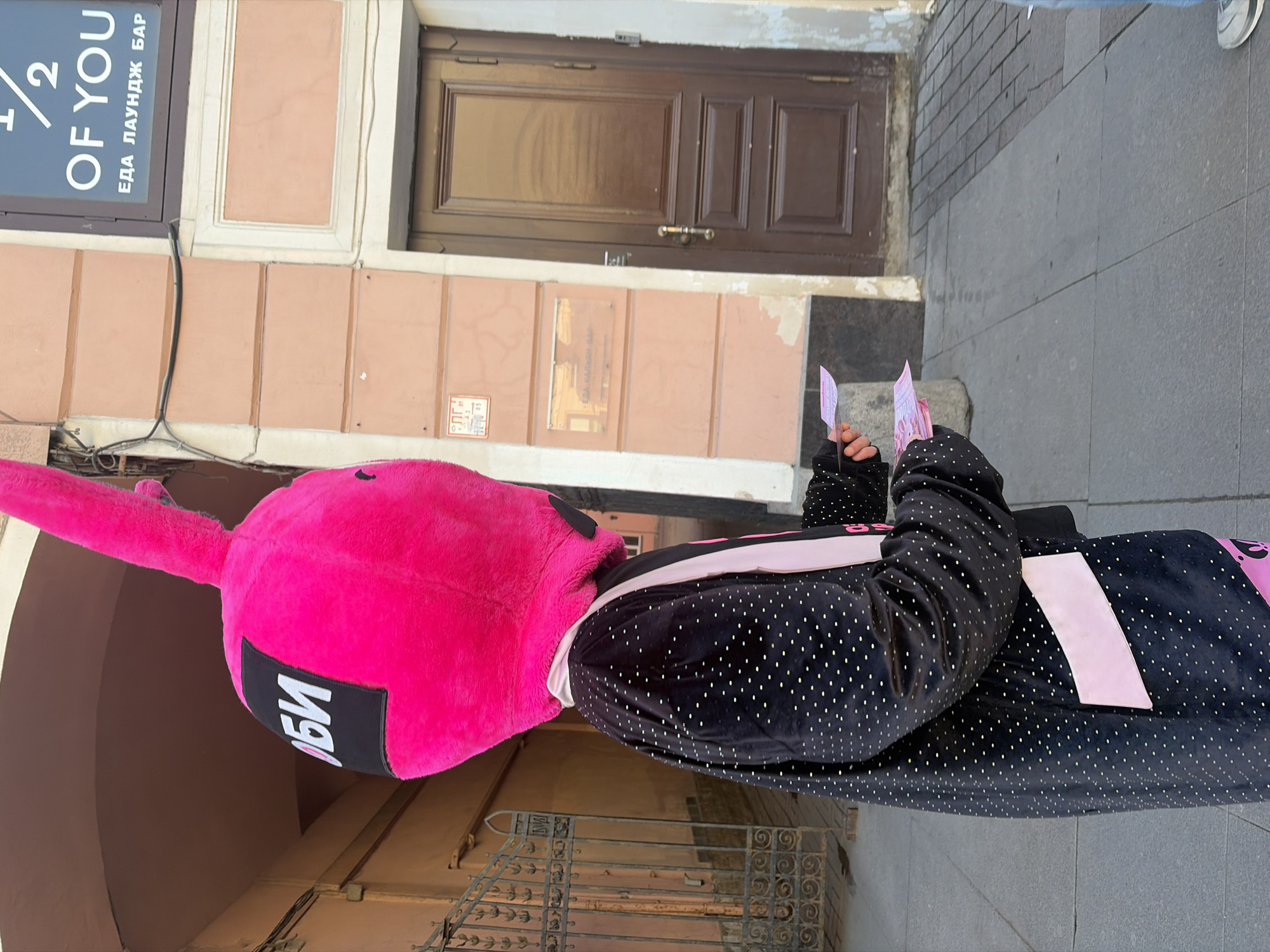 The image depicts a person in a bright pink, full-body bunny costume, standing outdoors on a street in Saint Petersburg, Russia. The costume features a black patch with white Cyrillic letters. The person is holding what appear to be pink leaflets or cards. They are dressed in a black, polka-dotted, velour tracksuit with pink stripes. The background includes a building facade, a brown door, a metal railing, and a sign that reads "1/2 OF YOU ЕДА ЛАУНДЖ БАР". The ground is paved with grey stone.