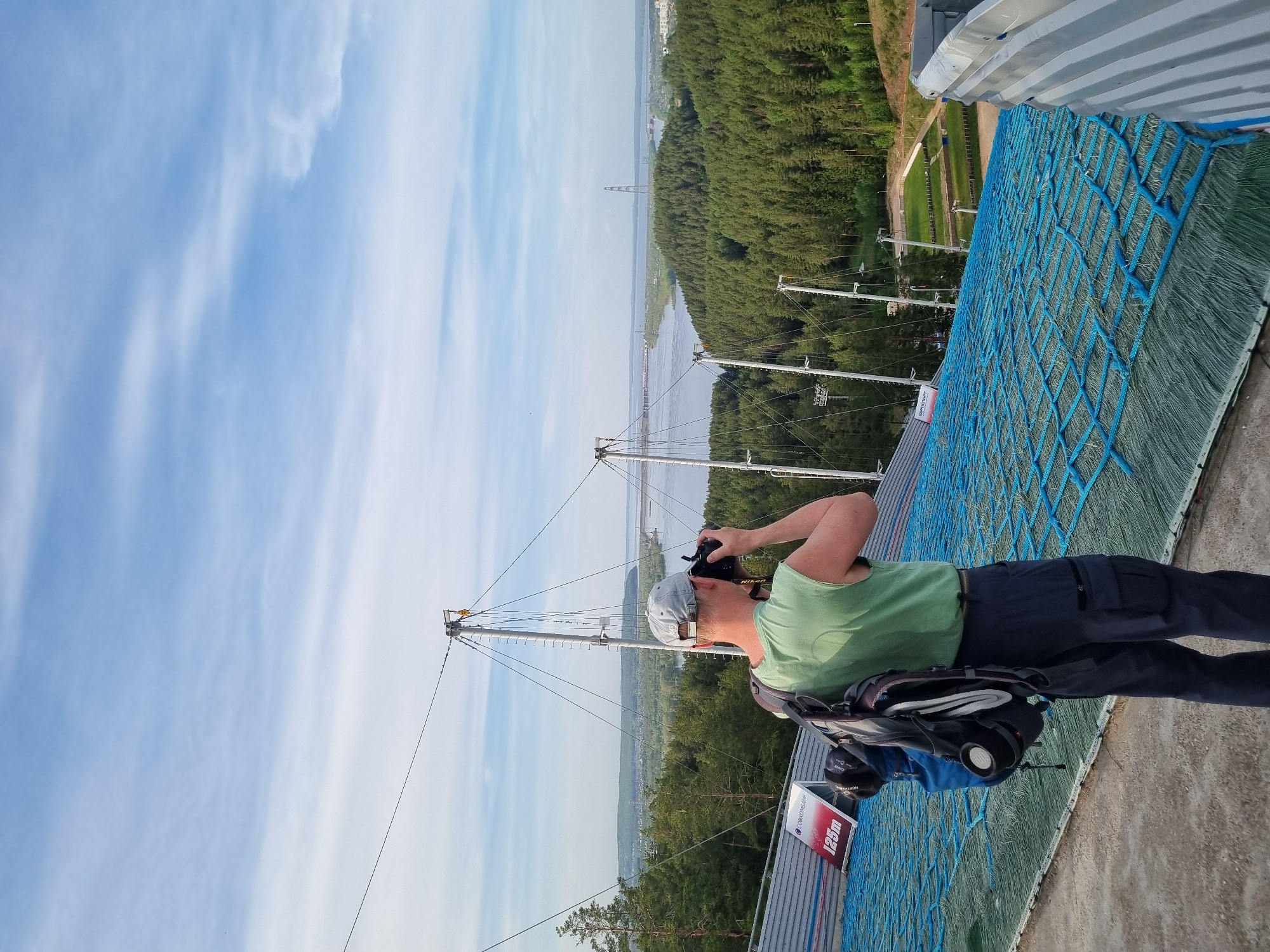 A man stands on a structure, possibly a ski jump, near the city of Прикамский, Russia. The man wears a green t-shirt, dark pants, a grey baseball cap, and has a backpack. He holds a camera to his face, appearing to take a picture of the view. The background shows a river, a forest, and some industrial structures. The structure the man stands on is covered with blue netting. A sign reading "125m" is visible. The sky is partly cloudy.