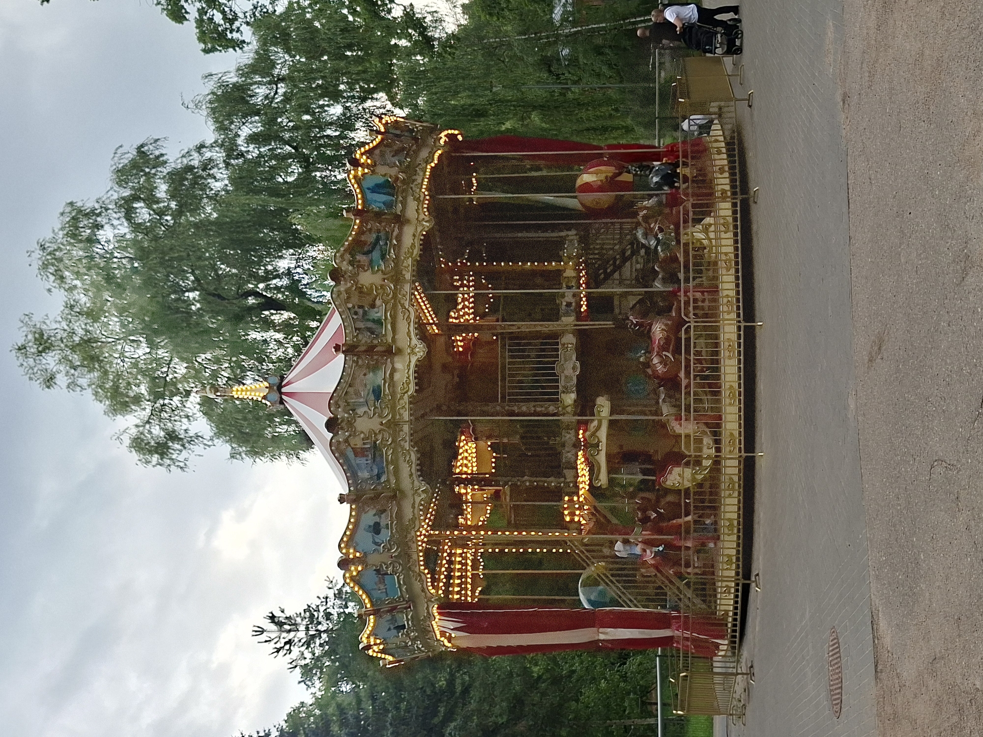 The image depicts a merry-go-round in a park setting, with lush green trees forming the backdrop. The merry-go-round is ornate, with a red and white striped roof, adorned with numerous golden lights. Various carousel animals, including horses, are visible inside. A few people are present; one child is seen on the merry-go-round, and two adults with a stroller are near the edge of the frame. A golden metal fence surrounds the merry-go-round. The ground is paved with interlocking bricks and asphalt. The sky is overcast, suggesting a cloudy day. This scene is likely located in Kaunas, Lithuania, in an amusement park or recreational area.