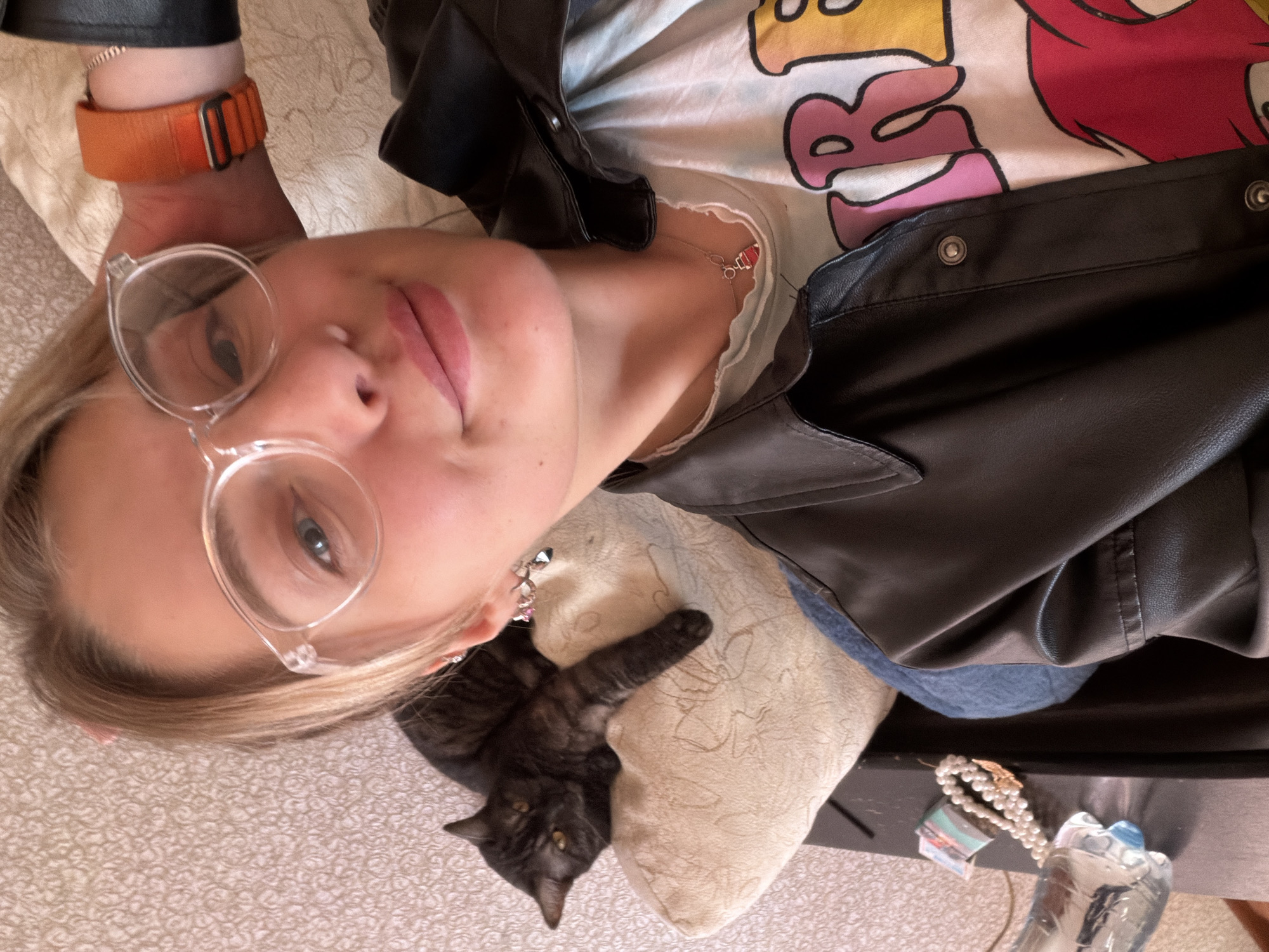 A woman wearing clear-framed glasses, a black leather jacket over a graphic t-shirt, and an orange Apple Watch band is lying on her side. A dark tabby cat is resting on a beige pillow near her. A strand of pearls and a water bottle are visible on a dark surface near the pillow. The location is suggested to be Naberezhnye Chelny, Russia.