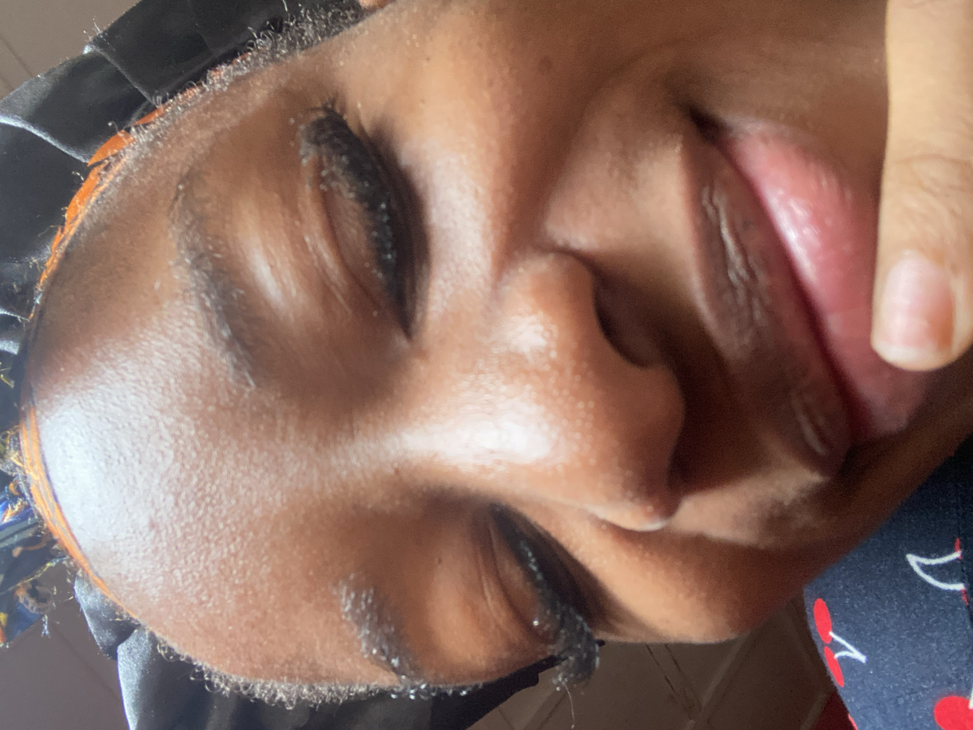 A close-up, low-angle portrait focuses on the face of an individual with a dark complexion, from the forehead to the chin. The individual's eyes are closed, featuring prominent, long, dark eyelashes. Dark, defined eyebrows are visible above the closed eyelids. The nose is centered, with visible nostrils. The mouth is slightly open, revealing part of the lower lip, which appears full and slightly textured, and some interior structures like the lower teeth or inner lip. A finger is positioned against the lower lip or chin area. The individual wears a dark-colored head covering, possibly satin or silk, with a band of orange fabric visible underneath it, near the hairline. A dark blue garment patterned with red and white cherries is visible in the lower left portion of the image. The background is highly blurred and indistinct, consistent with an indoor setting under ambient lighting. No discernible visual cues within the image indicate the specific location in the city of Jos, Nigeria.