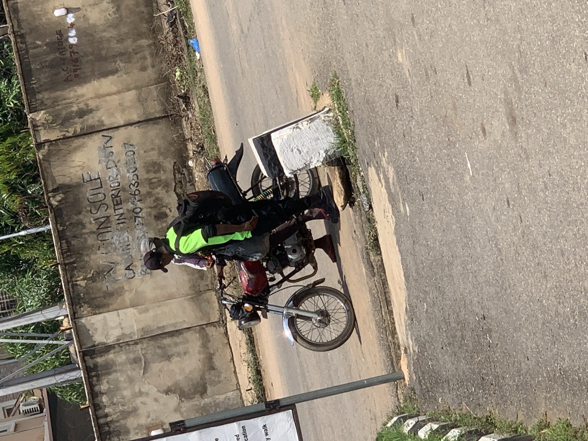 In Abuja, Nigeria, two individuals stand near a motorcycle parked on a paved surface next to a concrete wall. The person on the right wears a neon green vest over dark clothing and a black backpack. The person on the left, partially obscured, wears a white shirt and a dark cap. The motorcycle is a red commuter bike. The concrete wall behind them is marked with graffiti and some signs, including the words "TV CONSOLE," "INTERIOR DSTV," and a phone number that appears to be "270-46330207." To the far right of the wall, red spray paint reads "A.C. FALOCE" and a phone number "071679864." The environment appears to be an outdoor urban setting with some vegetation visible.