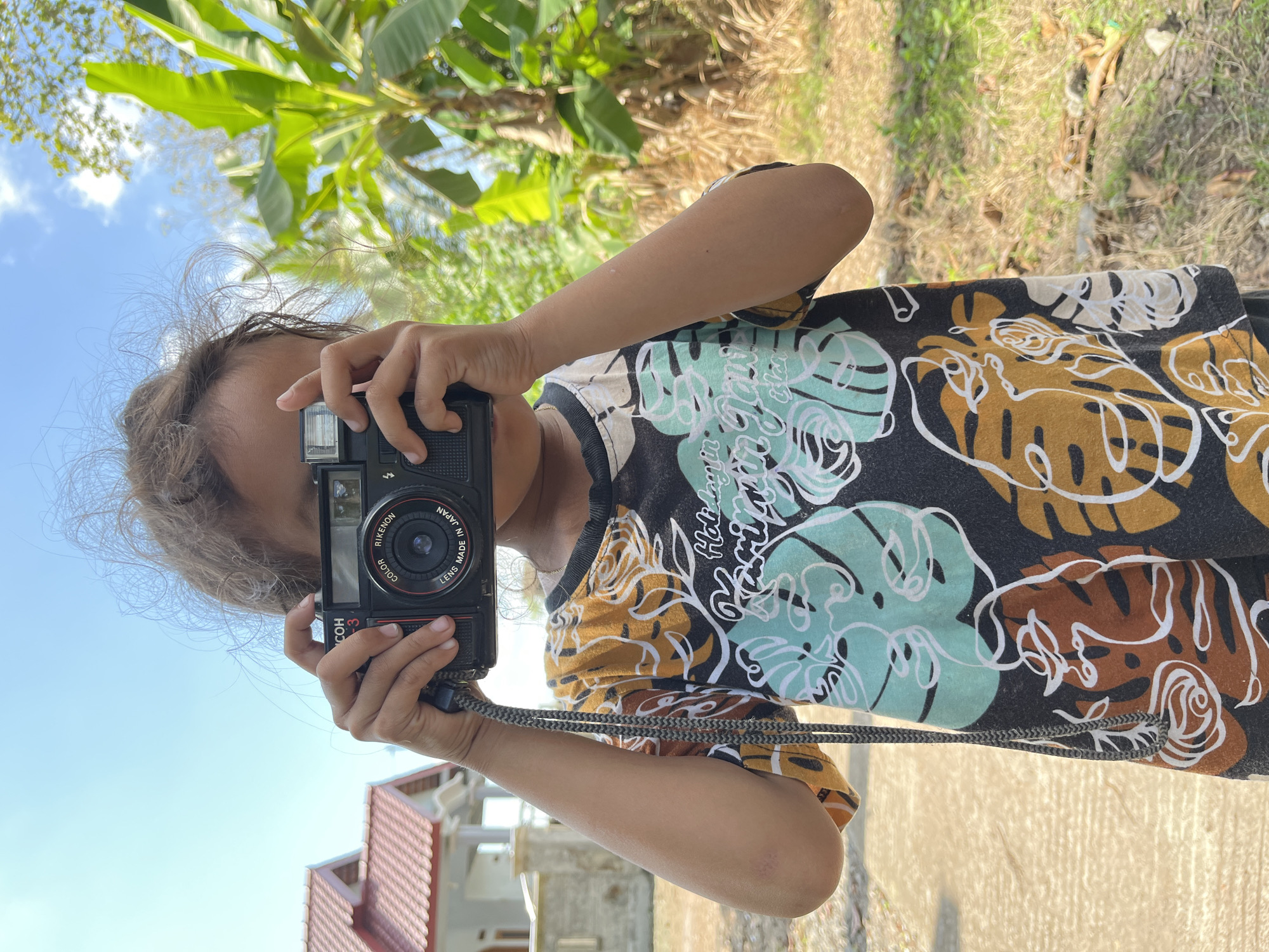 A young person is holding a black Ricoh camera to their face, obscuring most of their face, and looking through the viewfinder. The person is wearing a dark t-shirt with a colorful abstract pattern of tropical leaves and flowers in shades of teal, brown, and yellow. They are standing outdoors in what appears to be a residential area in Tambakreja, Indonesia, with a house with a red tiled roof visible in the background to the left. Lush green banana trees and other foliage are present to the right. The ground is a mixture of concrete and dry grass. The sky is a clear blue with a few wispy clouds. The lighting suggests it is daytime.