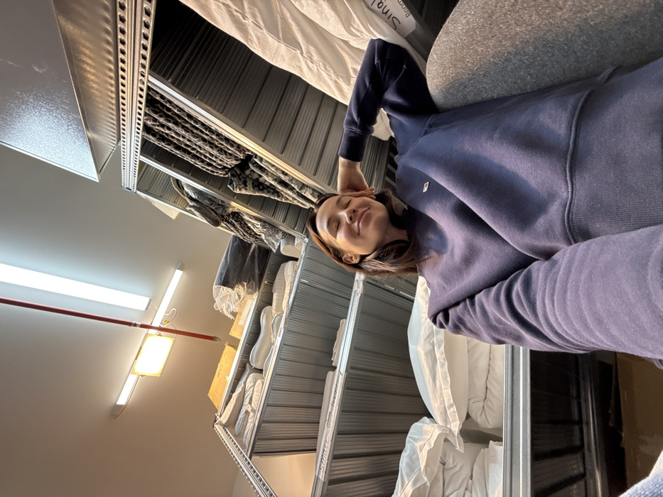 The image depicts a female individual within an indoor commercial storage or retail backroom environment. The subject, a female with dark brown hair, is shown from the chest up, angled upward, wearing a navy blue crew-neck sweatshirt with a small emblem on the left chest and grey pants. Her eyes are closed, and she exhibits a gentle smile, with her right hand positioned behind her head, indicating a relaxed posture. She appears to be leaning against a stack of white textiles in the foreground.
The background features tall, galvanized metal shelving units densely packed with merchandise. On the left, shelves contain numerous white pillows and folded textiles, some with visible labels including "Smart" and "Jeans." On the right, shelves hold folded or rolled patterned fabrics (resembling animal print) and white bedding, with one label partially visible as "Single / Room." The ceiling features multiple fluorescent strip lights and a central rectangular warm-toned light, with a red pipe running across it.
There are no visual cues within the image, such as specific signage, unique architecture, or local branding, to precisely identify a location within Tel-Aviv, Israel. The scene presents a generic interior typical of a textile or home goods warehouse or storage facility.