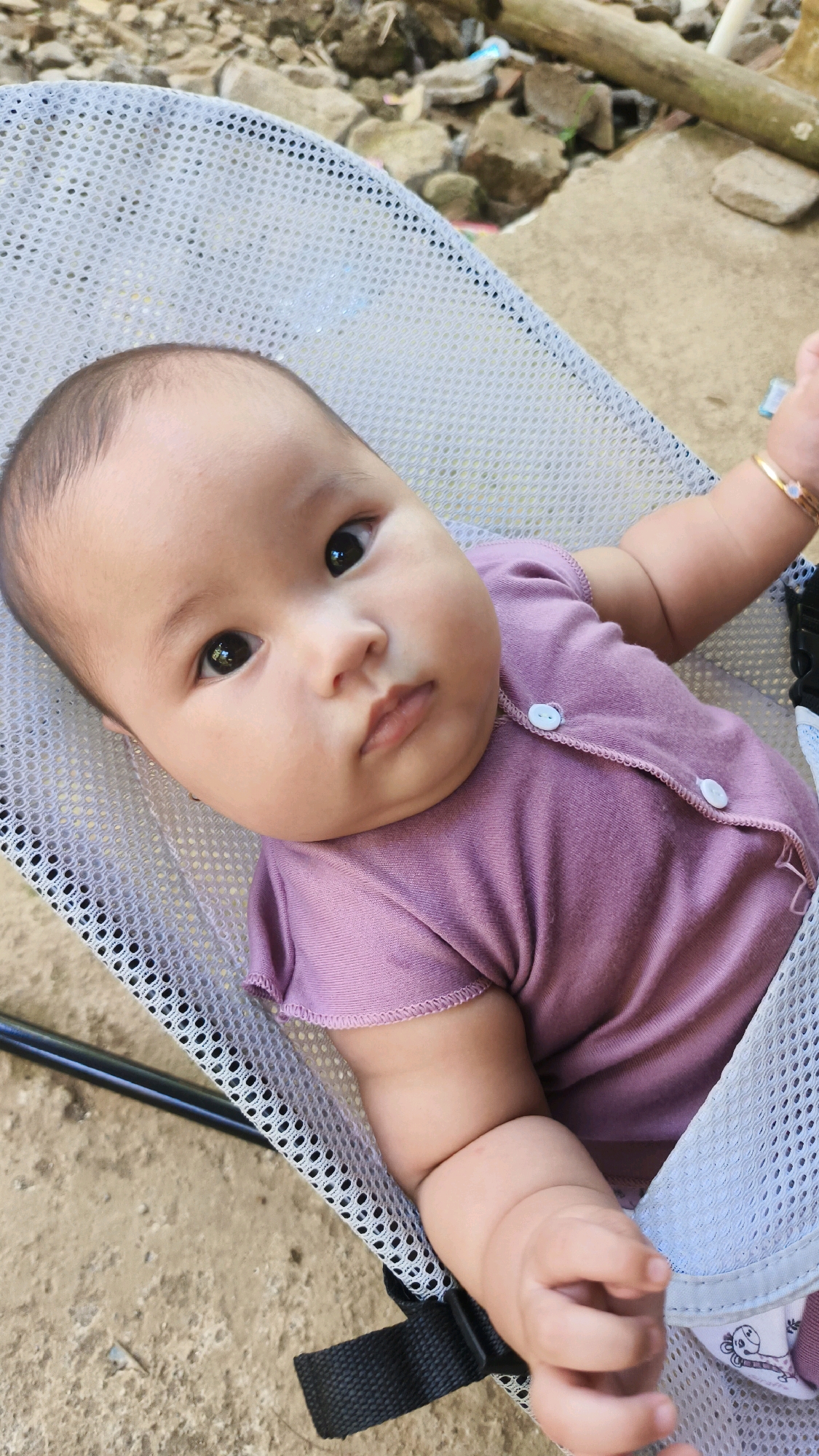 A baby girl with dark hair, large dark eyes, and chubby cheeks lies in a mesh baby bouncer. She is wearing a short-sleeved, light purple onesie with two white buttons on the front. The baby has a gold bracelet on her left wrist with a light blue stone. The bouncer is positioned on a sandy, outdoor surface with rocks and debris in the background. The scene appears to be located in Donorejo, Indonesia.
