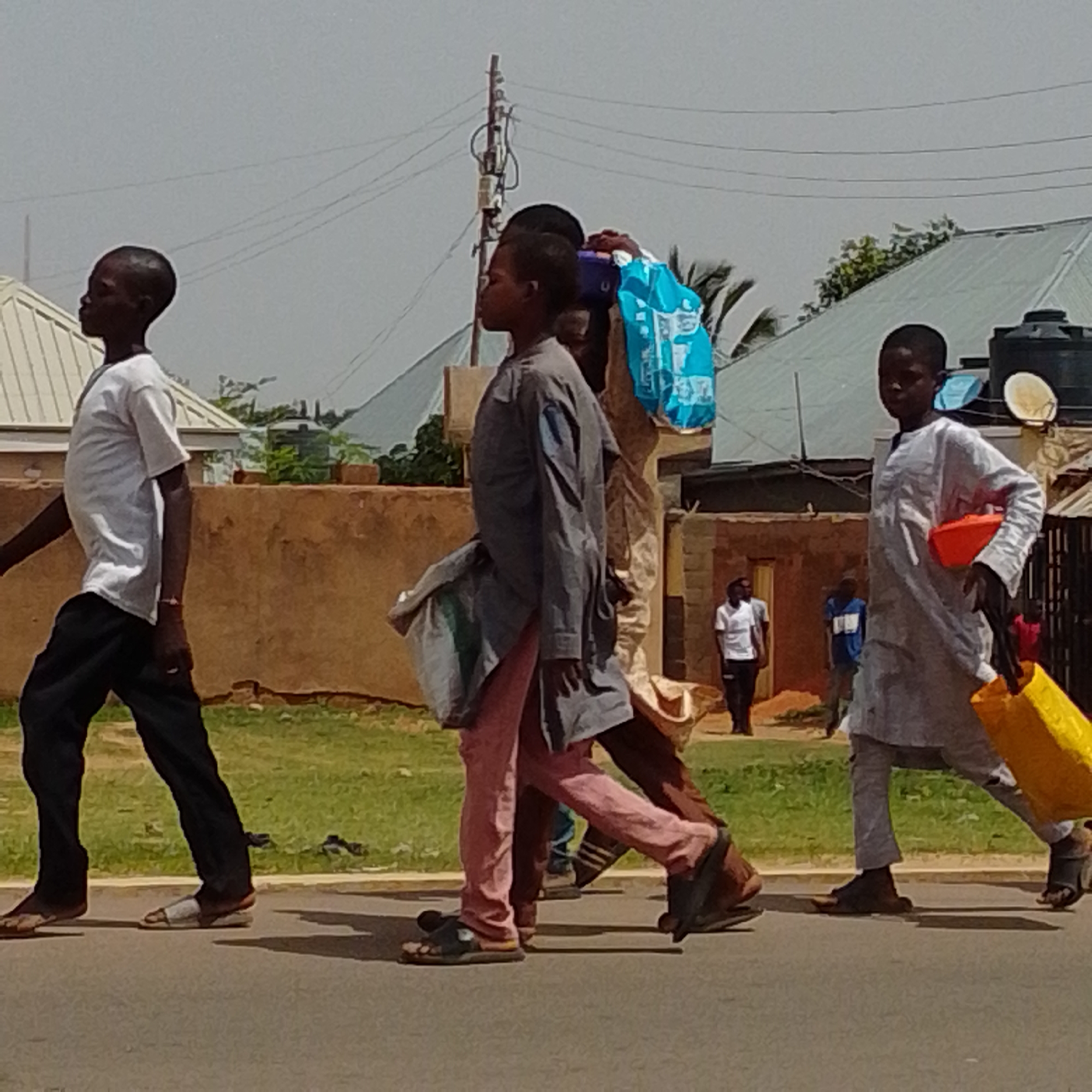 The image shows a street scene in Gombe, Nigeria, with several people walking on a paved road. In the foreground, a young boy wearing a white t-shirt and black trousers is walking from left to right, looking to his left. Next to him, a person wearing a gray long-sleeved shirt and pink trousers is walking in the same direction, carrying a bag over their shoulder. Behind them, another person in a dark shirt and trousers is walking. Further to the right, another young boy in a light gray traditional outfit is walking and carrying a bright orange object and a yellow bag. In the background, there are buildings with corrugated metal roofs, a wall, and some vegetation. A utility pole with wires is visible in the upper part of the image. The sky is overcast. The overall impression is that of a busy street with people engaged in everyday activities.