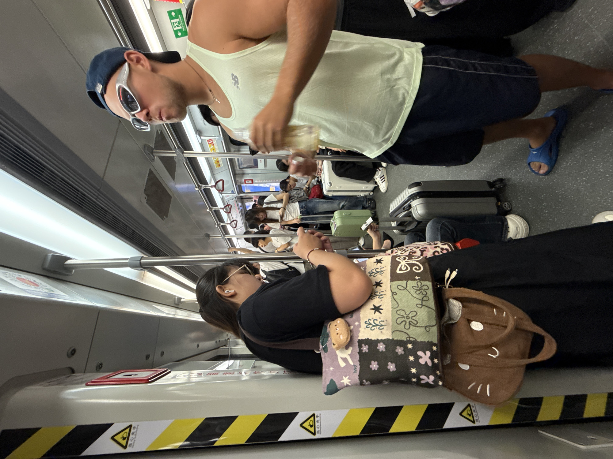 In Haizhu District, China, a man wearing a navy blue baseball cap, white wraparound sunglasses, and a light green sleeveless athletic shirt is holding a clear plastic cup with a yellow liquid. He is also wearing navy blue shorts and blue slip-on sandals. To his left, a woman with her hair pulled back is wearing a black t-shirt and glasses. She is holding a smartphone in her right hand and has two tote bags hanging from her left shoulder. One bag is a patchwork design with flowers and a small brown bear keychain, and the other is a brown bag with a cat face design. They are standing on a subway train. Other passengers and luggage, including suitcases, are visible in the background. The train car interior features grey walls, yellow and black striped safety markings, metal poles, and overhead lighting.