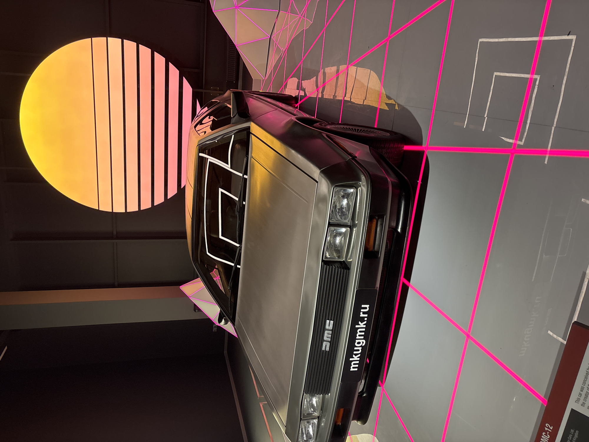 The image depicts a DeLorean DMC-12 car, a retro-futuristic sports car, displayed in a modern exhibition space. The car is positioned on a reflective surface overlaid with a neon pink grid pattern. In the background, a large, circular graphic resembling a sunset with horizontal lines radiates yellow and pink hues. Geometric, illuminated structures are visible on the right side of the car. A sign with the text "mkugmk.ru" is affixed to the front of the vehicle. The scene is dimly lit, with the neon lights and the sunset graphic providing the primary illumination. There are no people visible in the image. The exhibition appears to be dedicated to automobiles or a related theme, given the prominent display of the DeLorean. This exhibition is located in the Museum Complex of Military Technology in Verkhnyaya Pyshma, Russia.