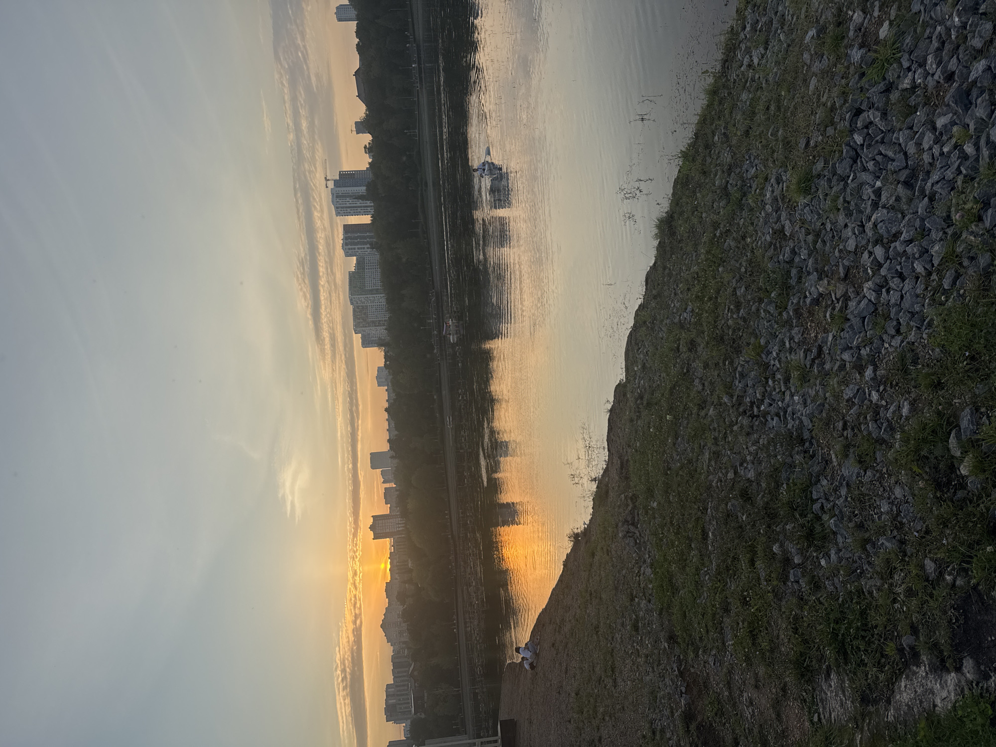 The image captures a serene view of the Belaya River in Ufa, Russia, during a sunset. The sky is painted with soft hues of orange and yellow as the sun dips below the horizon, casting a warm reflection on the water's surface. Tall buildings and trees form a picturesque backdrop along the riverbank. In the foreground, a rocky embankment slopes down to the water, with patches of green grass adding a touch of nature. Two individuals are seen near the river's edge, seemingly enjoying the tranquil atmosphere. Further out on the river, a lone figure rows a small boat, adding a sense of peaceful solitude to the scene. The overall impression is one of calm and natural beauty within an urban setting.