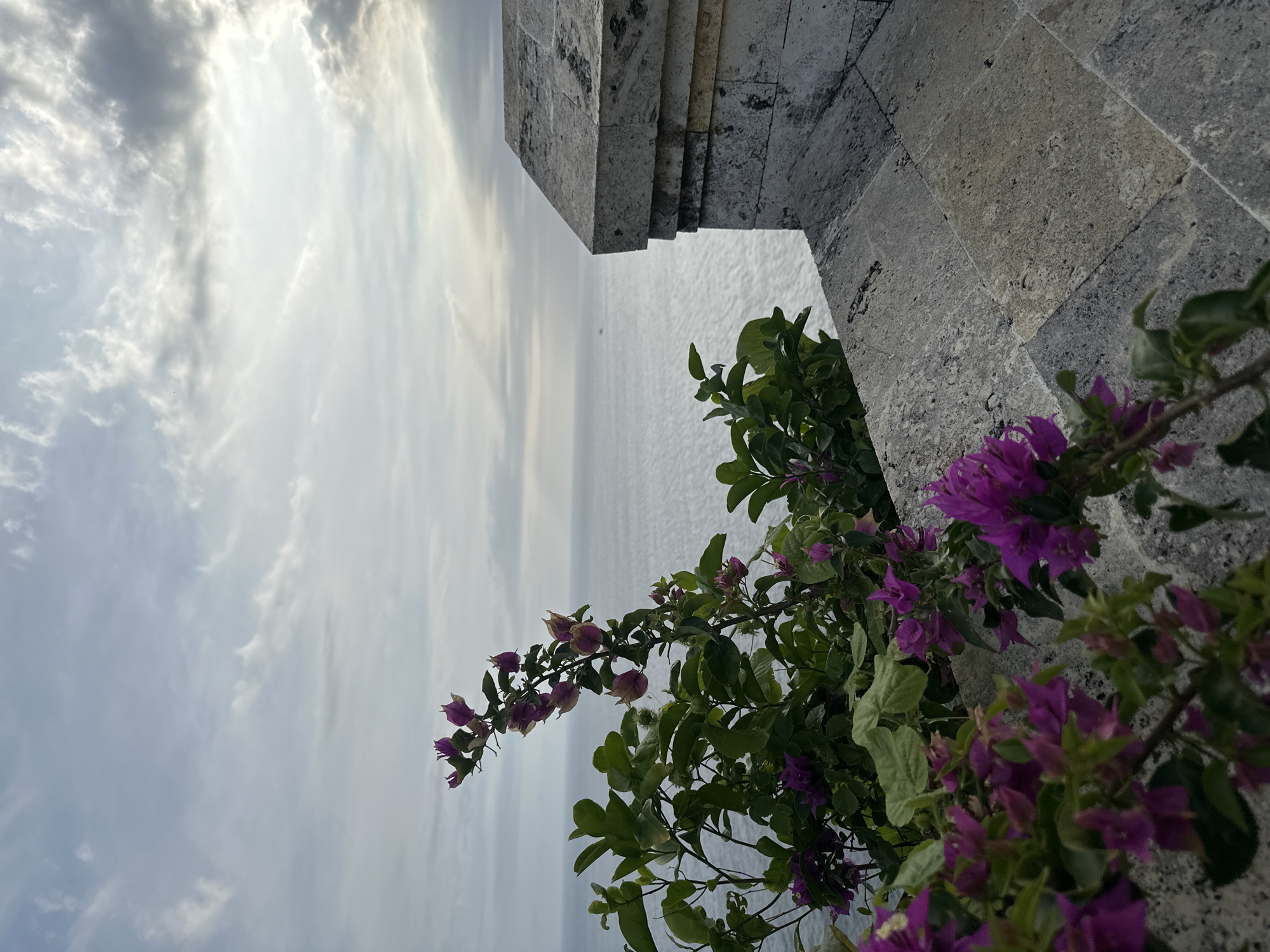 The image depicts a cloudy sky and ocean view from what appears to be a stone structure or wall adorned with bougainvillea flowers in Kuta Selatan, Indonesia. The stone structure is a light gray color with a layered design. The bougainvillea is a mix of green leaves and vibrant purple blossoms. The ocean is calm and light gray, mirroring the sky above.