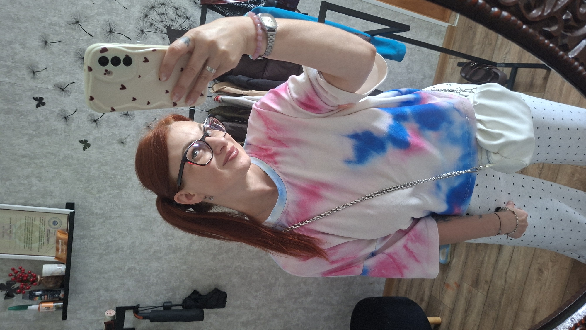A woman with auburn hair pulled into a ponytail and wearing glasses is taking a mirror selfie. She is wearing a tie-dye t-shirt with pink, blue, and white patterns, and grey polka dot pants. She has a silver chain bag slung across her body and a white pouch bag on her hip. She is wearing a watch on her left wrist and rings on her fingers. The background shows a wall with a dandelion decal, a shelf with decorative items, and a clothing rack with jackets. The floor is wooden. The scene appears to be indoors, possibly in a home in Myadzyel, Belarus.