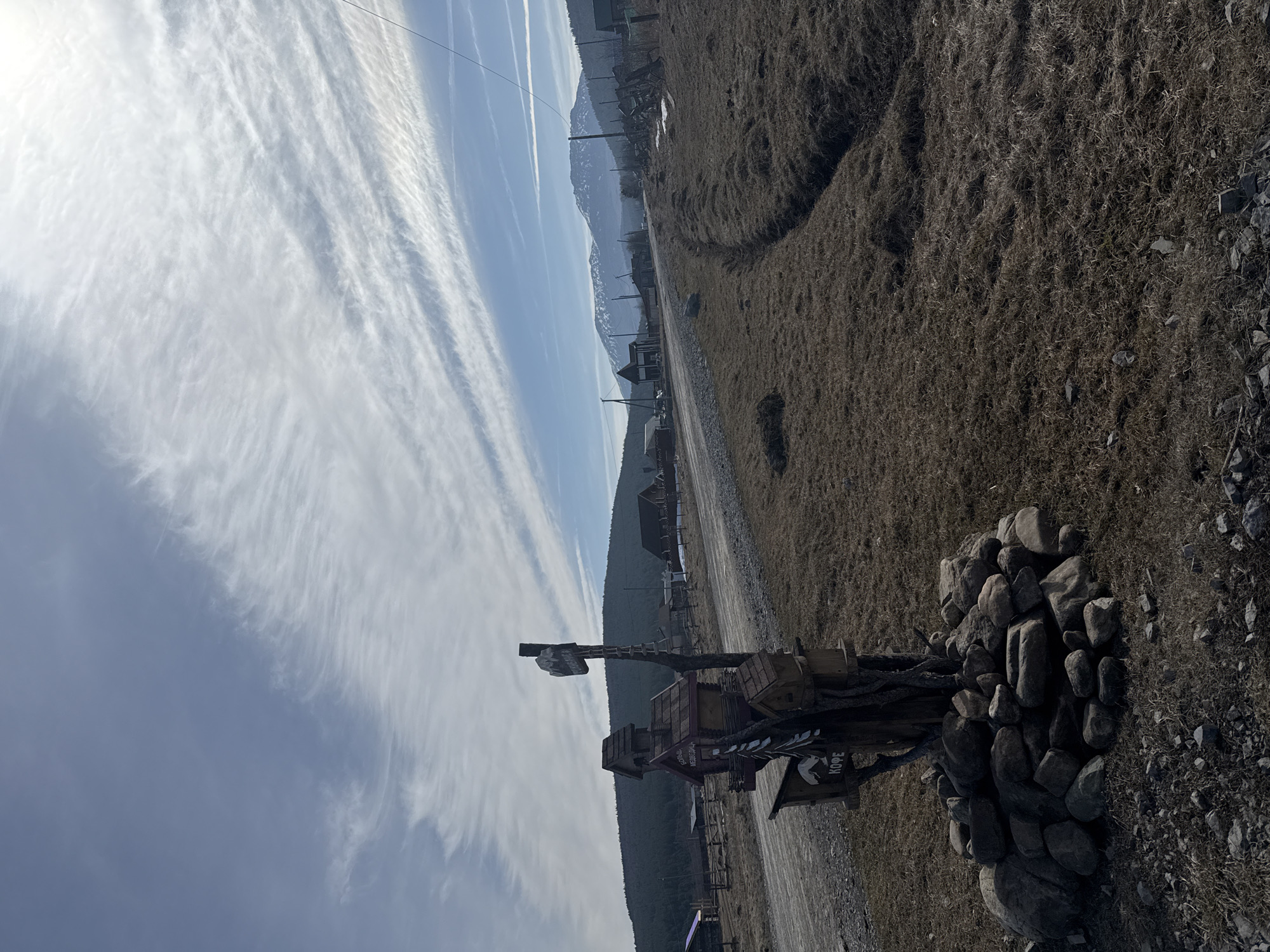 The image shows an outdoor scene in Tyulyuk, Russia, featuring a partly cloudy sky with streaky clouds. A rural road leads into the distance, flanked by dry vegetation. On the right, a pile of rocks surrounds a wooden structure with multiple birdhouse-like sections and signboards with text in Russian. In the background, there are buildings and trees, and a mountain range is visible on the horizon. Wires are visible against the sky.