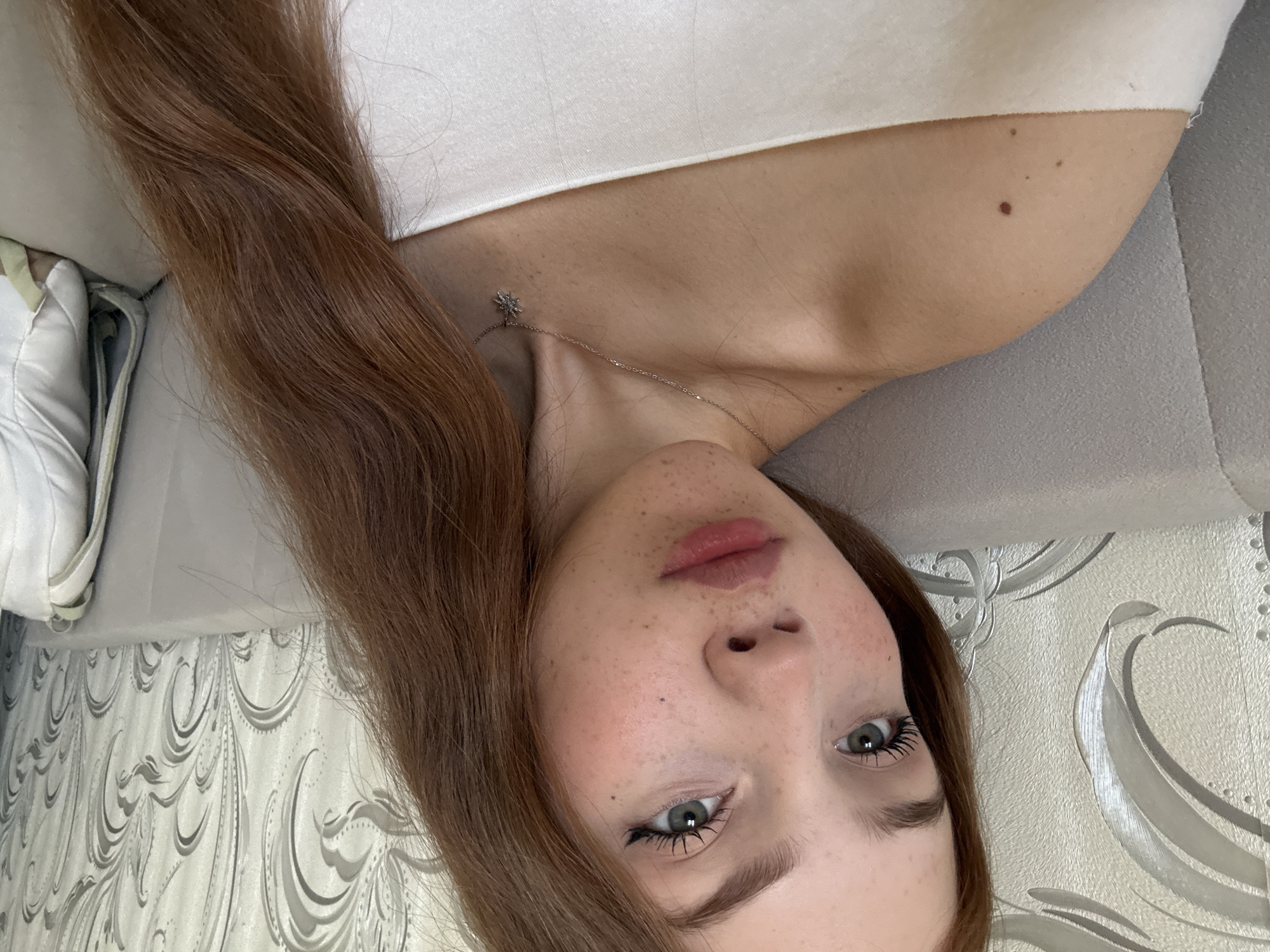 The image presents a close-up portrait of a young woman lying down in an indoor setting, likely in Naberezhnye Chelny, Russia. She has long, wavy reddish-brown hair that frames her face and flows over her shoulders. Her skin is fair, notable for a scattering of freckles across her nose and cheeks, and several small moles visible on her neck and shoulder. Her striking green eyes look directly at the camera, with visible mascara enhancing her eyelashes. Her lips are full with a natural, soft pink tint.
She is wearing a simple white top, and a delicate silver chain necklace with a small, star-shaped pendant rests on her collarbone. The woman is positioned on a light grey, upholstered surface, possibly a couch or bed. In the upper left corner, a white sneaker is partially visible. The background reveals a wall covered with patterned wallpaper featuring intricate silver and white swirling designs. The overall atmosphere is relaxed and candid, with the woman displaying a neutral to slightly pensive expression. There is no visible text in the image.