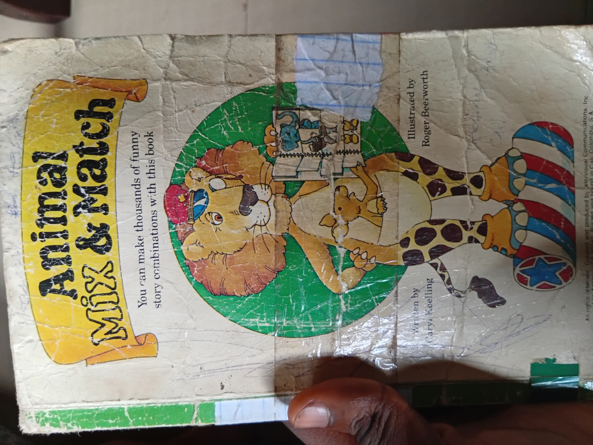 The image is a close-up of the cover of a children's book titled "Animal Mix & Match". The book cover is worn and creased. A cartoon lion with a crown is the central illustration, wearing a giraffe's body and large, striped boots. The lion is holding a smaller kangaroo-like creature in its pouch and another illustration of various animals in a grid-like format is visible on its chest. The book states "You can make thousands of funny story combinations with this book". The author is listed as Caryi Koelling and the illustrator as Roger Beerworth. The image is captured from an eye-level perspective and the lighting appears to be natural, casting some soft shadows. A person's hand is partially visible on the left side of the frame, suggesting the book is being held. The background is indistinct and appears to be an indoor setting, possibly a room in Nkpor, Nigeria, though no specific city landmarks are visible.