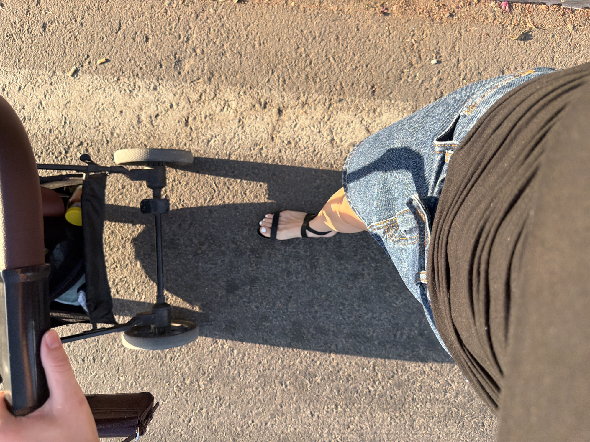 The image shows a first-person perspective of someone walking with a stroller on a paved surface. The subjects are a person's legs and feet, a stroller, and the ground. The person is wearing blue denim shorts and black sandals with white toenail polish. The stroller is partially visible, with one wheel and part of its frame and fabric. The ground appears to be asphalt or a similar paving material, with some scattered debris, possibly cigarette butts.
The activity taking place is walking or pushing a stroller. The time of day appears to be late afternoon or early evening, indicated by the long shadows cast on the ground, suggesting the sun is low in the sky. The lighting is warm and golden. The setting is likely an outdoor public space, such as a sidewalk, park path, or promenade.
Notable details include the texture of the paved surface, the sunlight creating distinct shadows, and the individual's attire. The presence of cigarette butts could suggest a public area where smoking is permitted. The overall mood conveyed is casual and perhaps leisurely, given the context of walking with a stroller. There is no visible text in the image. The location context provided is Sharm El Sheikh, Egypt, which aligns with the warm lighting and outdoor setting.