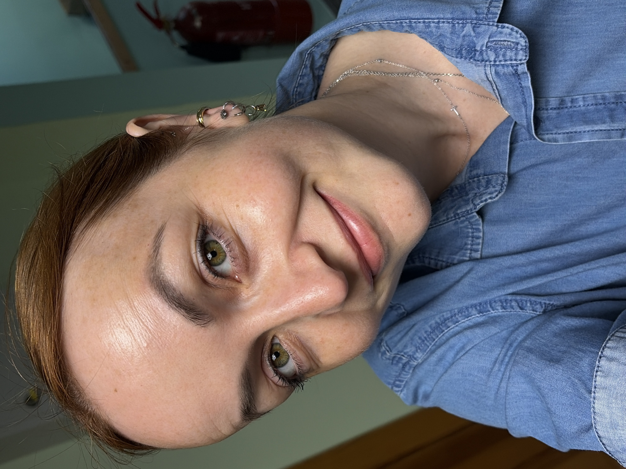 A close-up selfie shows a fair-skinned woman with reddish-brown hair and hazel eyes, looking directly at the camera. She is wearing a blue denim shirt and a silver necklace with a small cross pendant. Her face is slightly tilted, and she has a subtle smile. Multiple piercings are visible in her left ear. The background is blurred, but a green wall and a red fire extinguisher can be distinguished. The image does not contain any information that would allow for a determination of its location in Chaikovsky, Russia, or any specific city.