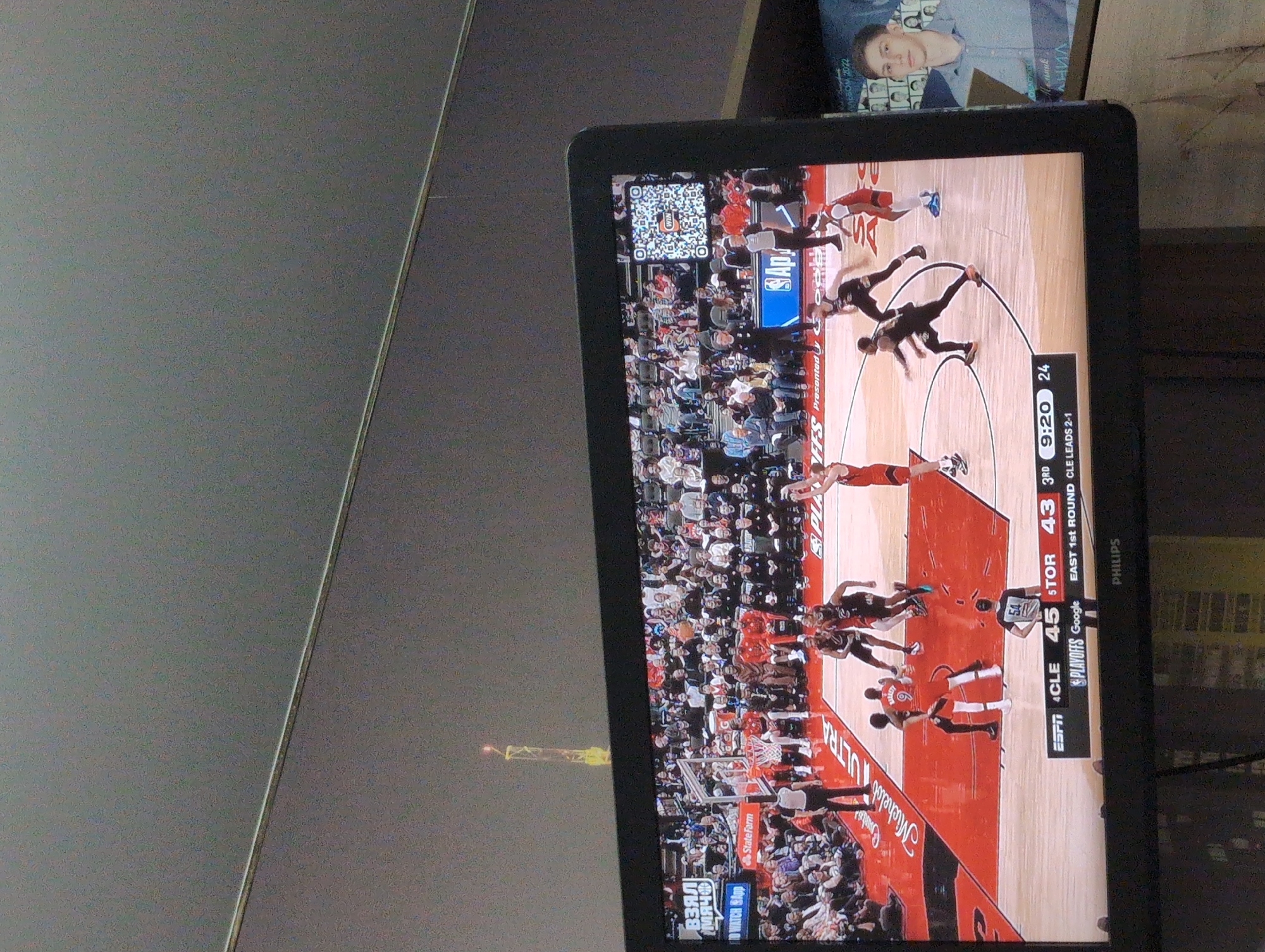 The image shows a basketball game being televised on a Philips monitor. The game is between the Cleveland Cavaliers (CLE) and the Toronto Raptors (TOR). The Cavaliers are leading 45 to 43 in the 3rd quarter with 9:20 on the clock. The game is the first round of the Eastern Conference playoffs, with Cleveland leading the series 2-1. The monitor also displays advertisements for Michelob Ultra, State Farm, and Google. In the upper right corner of the image is a photo of a young man. The background shows a gray tabletop. The photo was not taken in Izhevsk, Russia.