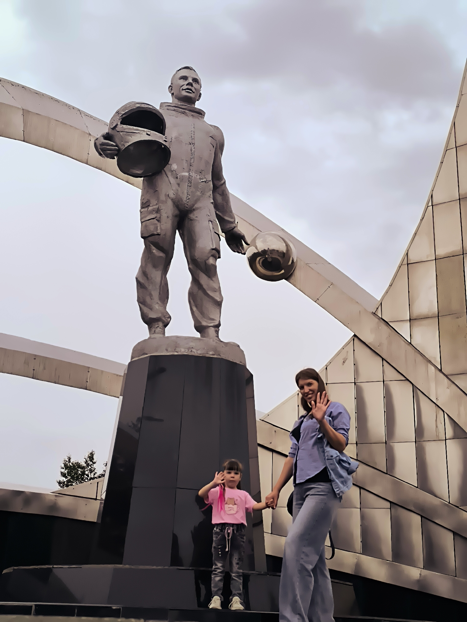 In the city of Qarağandı, Kazakhstan, a large bronze statue of a cosmonaut stands prominently, holding a helmet in one hand and a spherical object in the other. The statue is situated on a dark, multi-tiered pedestal. In front of the statue, two individuals are present: a young girl in a pink t-shirt and jeans, and an adult woman in a blue shirt and jeans. The girl is on the left, holding her right hand up. The woman is on the right, holding the girl's left hand and waving with her right hand. They are positioned on steps leading up to the monument. The background features a modern building with a metallic facade and an overcast sky.