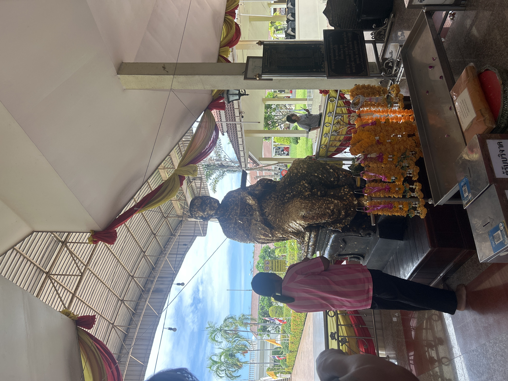 The image depicts an outdoor covered area in Thai Ko Subdistrict, Thailand, where a large, seated statue, adorned with gold leaf, is the central focus. A person wearing a pink and white striped shirt and black pants stands before the statue, facing away from the camera, with their hands clasped in front of them, suggesting a pose of prayer or reverence. In the background, palm trees and a building with a tiled roof are visible under a cloudy sky. Decorative drapes in red and yellow hang from the ceiling, adding to the ambiance of the location. To the right of the statue, a metal tray holds offerings, including several flower garlands and a folded orange cloth. A sign with Thai script is also visible on the right side of the image. Another person, wearing a grey shirt and sunglasses, is seen in the middle ground, also appearing to be observing the scene. The ground is a polished, reddish-brown material.