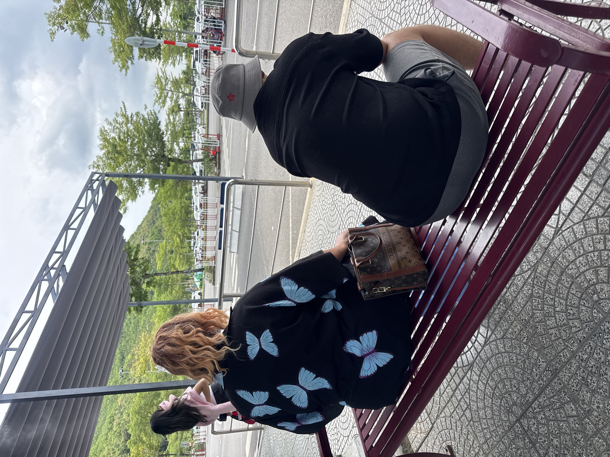Two people are sitting on a red bench next to a street. One person wears a black shirt, grey shorts, and a grey bucket hat with a red star. The other person wears a black shirt with blue butterflies and has a brown Louis Vuitton handbag on the bench next to them. The bench is on a sidewalk with a patterned ground. Behind the bench is a covered walkway, a road, a parking lot with cars, and some trees. The scene appears to be in the city of Đà Nẵng, Vietnam.