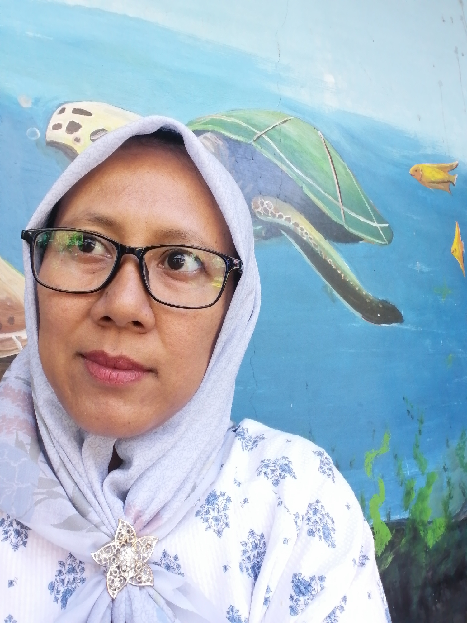 The image is a selfie of a woman wearing a light blue hijab and a white shirt with a blue floral pattern. She is also wearing a silver flower-shaped brooch on her hijab. She has dark eyes and is wearing black-rimmed glasses. The background is a mural of an underwater scene, with a sea turtle and fish painted on a blue wall. The woman is standing in front of the mural, which appears to be located outdoors. The overall scene suggests a casual and artistic environment.