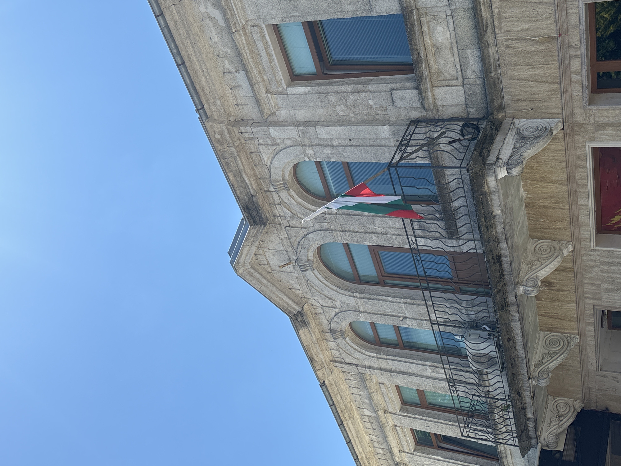 The image shows the exterior of a building in Kavarna, Bulgaria. The building is made of light-colored stone with arched windows. A balcony with a wrought-iron railing extends across a portion of the facade. A Bulgarian flag is displayed on the balcony. The sky is clear and blue. An air conditioning unit is visible on the balcony. The architecture suggests an older building, possibly a public building or a historic residence.