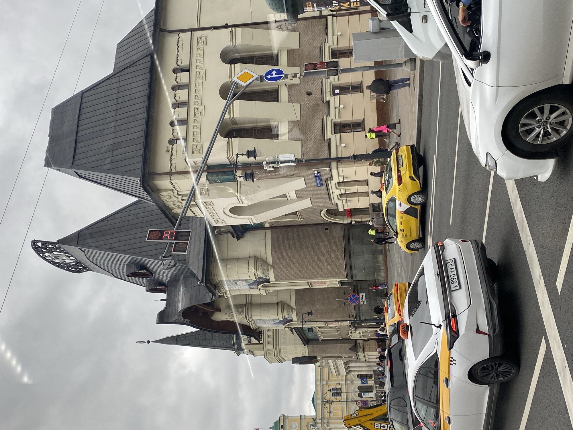 A street scene unfolds under an overcast sky in front of a large, ornate building with a dark, multi-peaked roof. A clock tower is visible in the distance to the left. The building features cream-colored walls with decorative molding and arched windows. Several yellow taxis, some with "Yandex Taxi" branding, are present on the street. One white taxi with yellow accents and Russian license plates is in the foreground, angled to the right. A construction crane with a yellow boom is parked on the left side of the street. Pedestrians are scattered on the sidewalk and crossing the street. Traffic lights and street signs are visible. Power lines crisscross the sky.