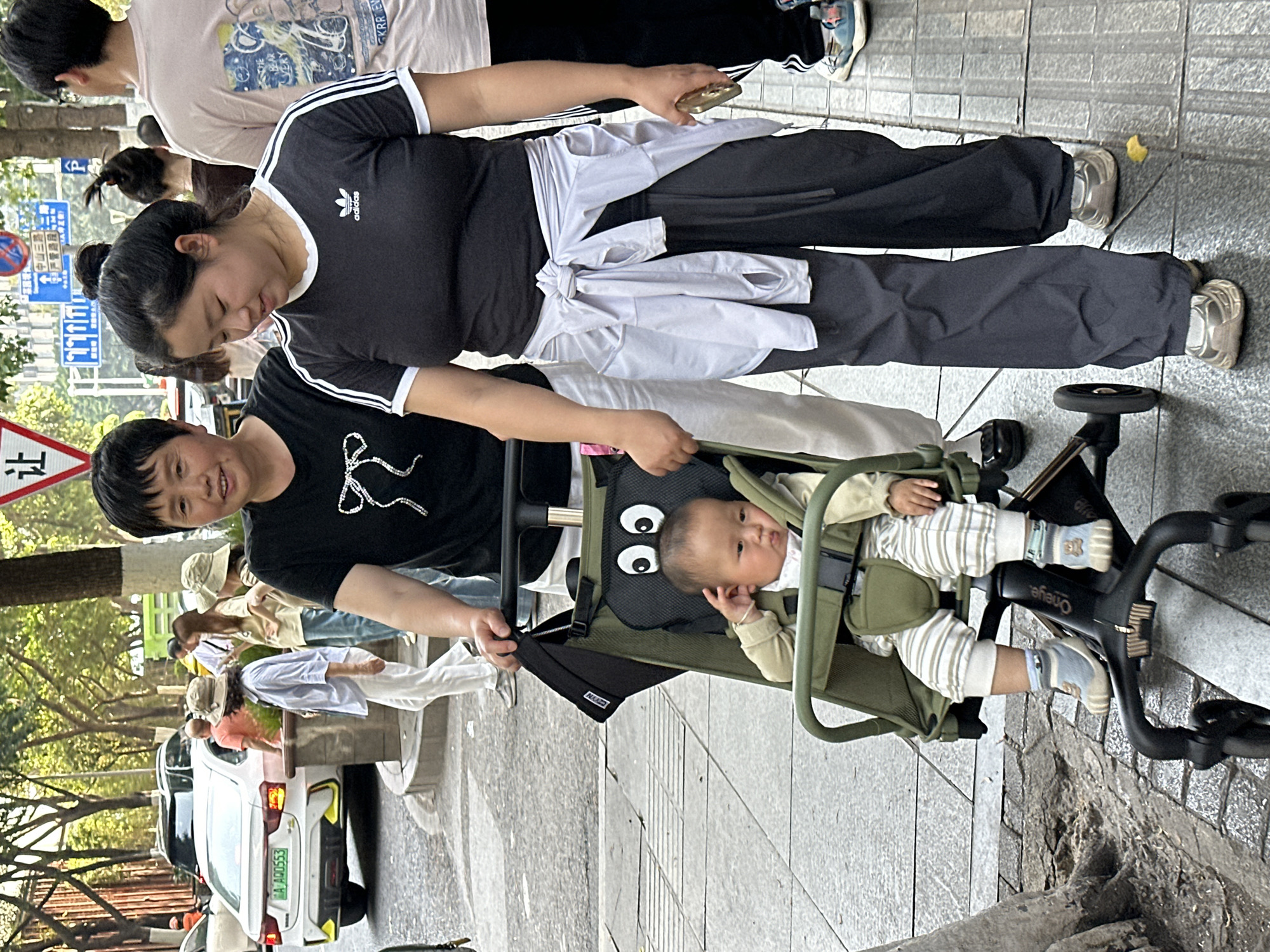 This image, rotated 90 degrees clockwise, captures a daytime street scene in what appears to be an urban area, likely Yuzhong District, China. The primary subjects are two adults and a baby in a stroller, walking on a paved sidewalk.
In the foreground, a woman with short dark hair, wearing a black t-shirt with a white design and dark pants, smiles towards the viewer as she pushes a green baby stroller. Seated in the stroller is a baby dressed in a light-colored striped outfit and patterned socks, looking calmly to the side. The stroller features a black cushion with a distinct white eye design and "Oneye" visible on its frame.
Behind the stroller, another woman, wearing a black Adidas t-shirt with white stripes, dark pants, and a white long-sleeved shirt tied around her waist, is looking downwards. She has dark hair styled in a bun and is holding a gold-colored smartphone in her right hand.
The background shows other pedestrians and a street with parked vehicles. A white car with a green license plate reading "A D10553" is partially visible. Prominent traffic signs include a red triangular "让" (yield) sign and several blue rectangular signs with white Chinese characters and symbols, indicating road directions or information. Green trees line the street, suggesting a pleasant outdoor environment. The lighting indicates it is daytime with good visibility.