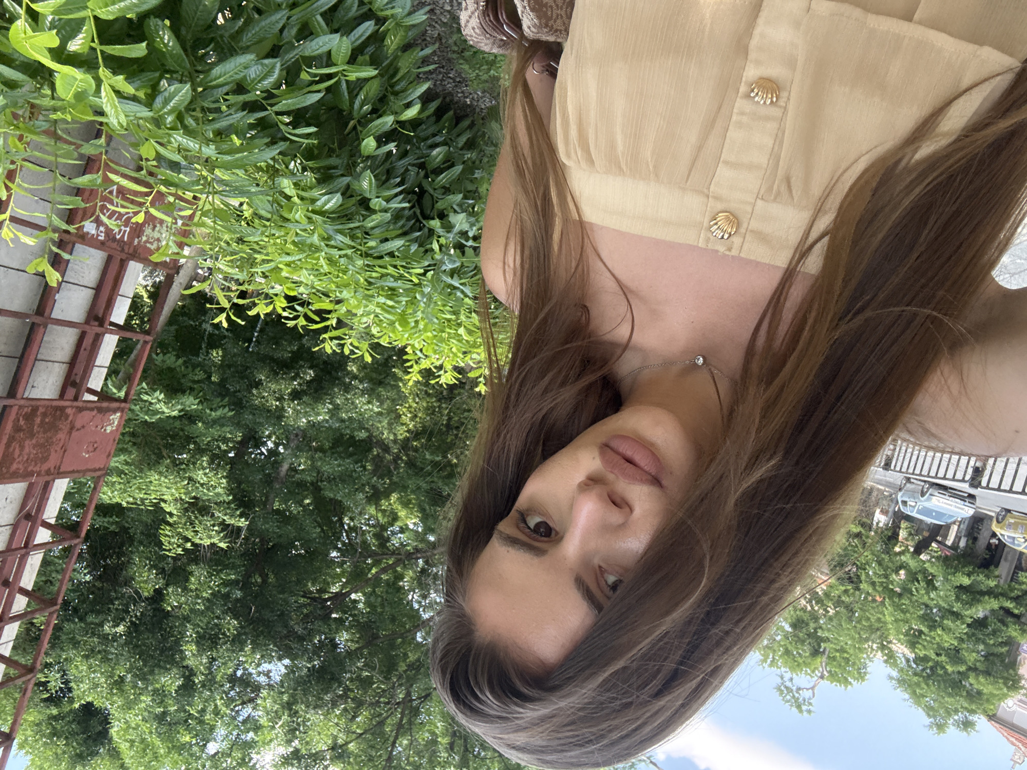 A young woman is featured in an outdoor portrait, likely a selfie, captured during daylight hours under clear skies. The image is angled, showing her looking directly at the camera with a neutral expression. She has long, dark brown hair and is wearing a light beige top with decorative gold shell-shaped buttons and a delicate silver necklace with a clear gem. A portion of a brown and beige patterned bag is visible on her right side.
The background is lush with vibrant green foliage from trees and bushes. A rusty red metal structure, possibly a fence or part of a building, is visible against a light-colored wall with faint, illegible markings. Further in the background, elements of an urban environment are discernible, including a street with parked cars (one light blue, one yellow) and distant buildings, all under a bright blue sky with scattered white clouds. The setting appears to be a green area within an urban landscape in Plovdiv, Bulgaria.