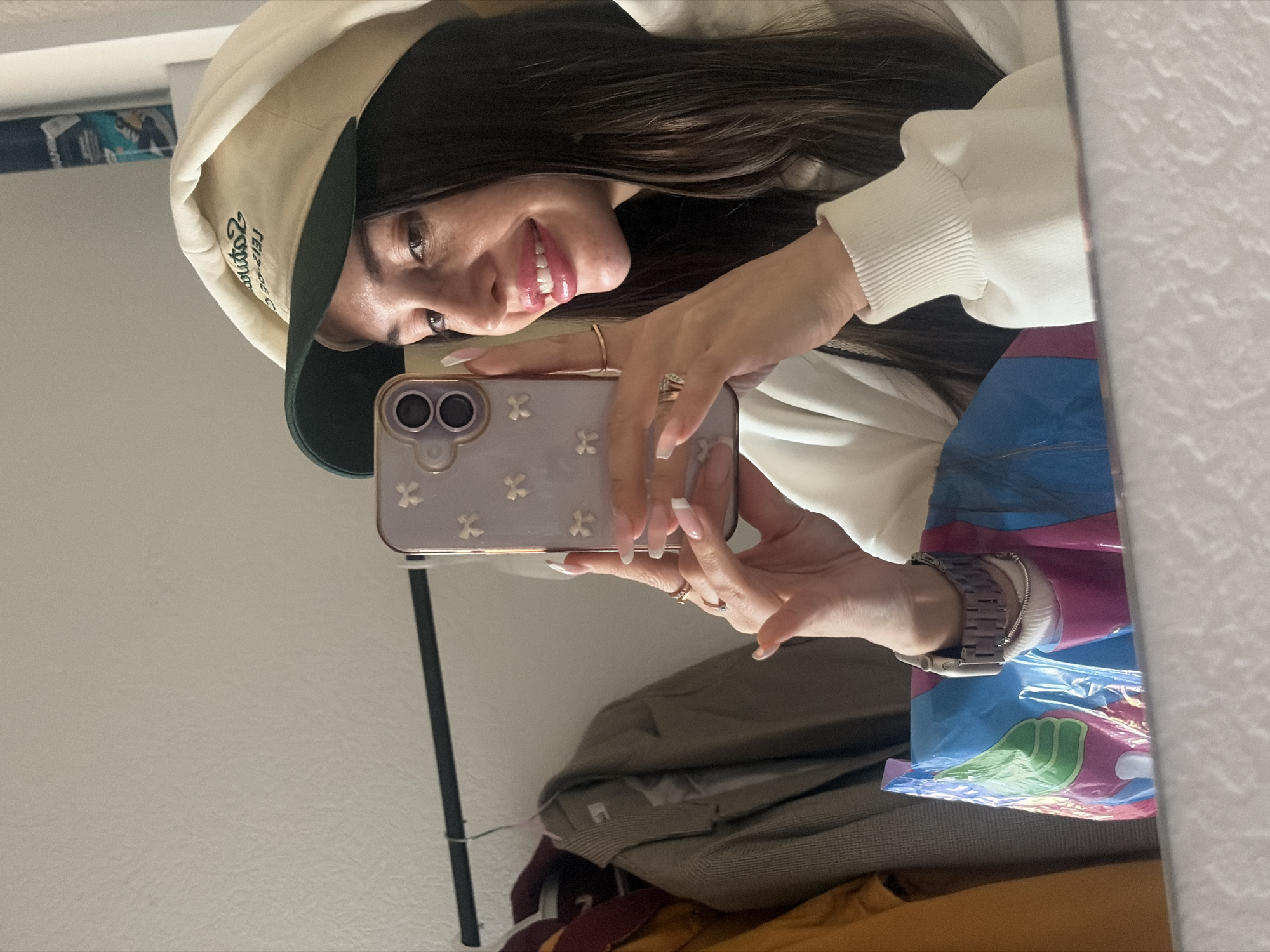 A woman with long dark hair and a beige cap is taking a selfie in a mirror. She is wearing a white sweatshirt and a watch on her left wrist. Her phone has a light purple case with white clover patterns. The background shows a white wall, a black bar (possibly part of the mirror or closet), and some clothing items. It is impossible to determine from this image if the location is in Saint Petersburg, Russia.