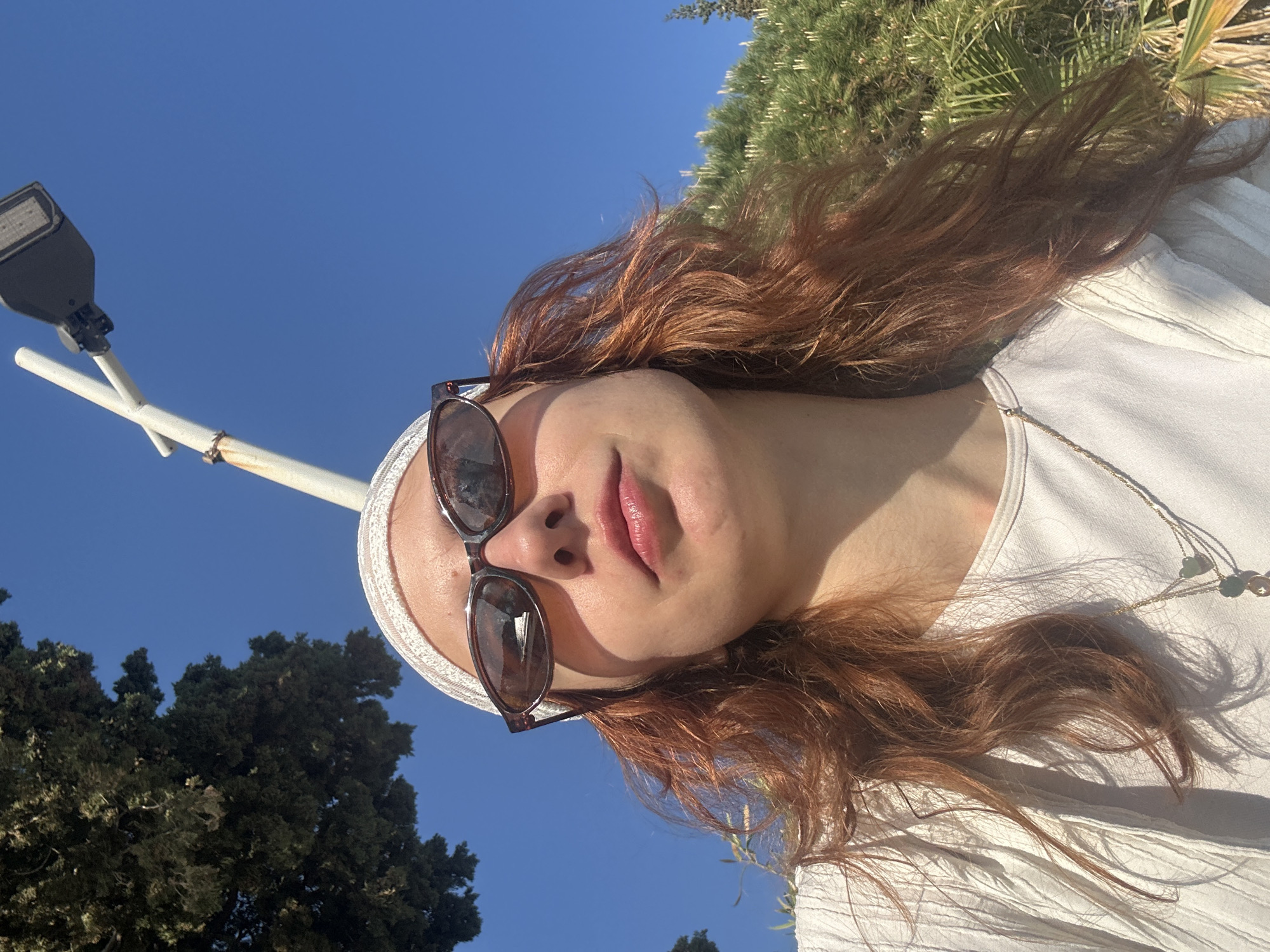 A person with reddish-brown hair and sunglasses stands outside under a clear blue sky, likely in Petrovac centar, Montenegro. A white bandana adorns their head, and they are wearing a light-colored, possibly white, top with a gold necklace. The person is positioned at an upward angle, looking down at the camera. To the left is a lamppost, and trees are visible in the background. Green foliage is present above and to the right of the subject.