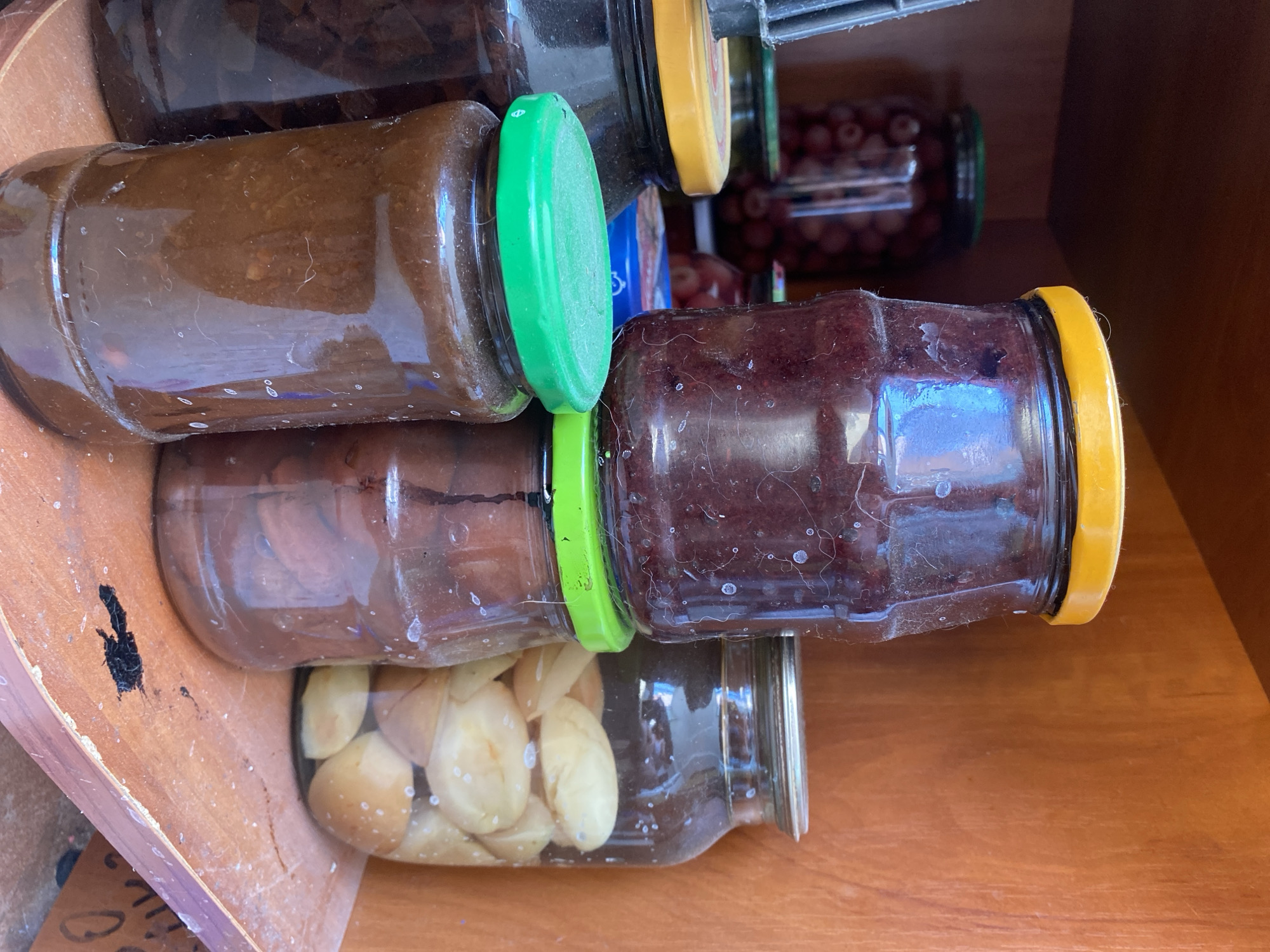 This image depicts a close-up view of several glass jars filled with preserved food items, likely jams or pickled fruits, stored on a wooden shelf. The jars have various colored lids: yellow, green, and silver. In the background, more jars containing red berries are visible. The wooden shelf appears to be part of an indoor storage area, possibly a pantry or a cellar, given the lighting and the presence of multiple preserved food containers. The overall impression is one of home preservation and storage. The location is Chornomorsk, Ukraine.