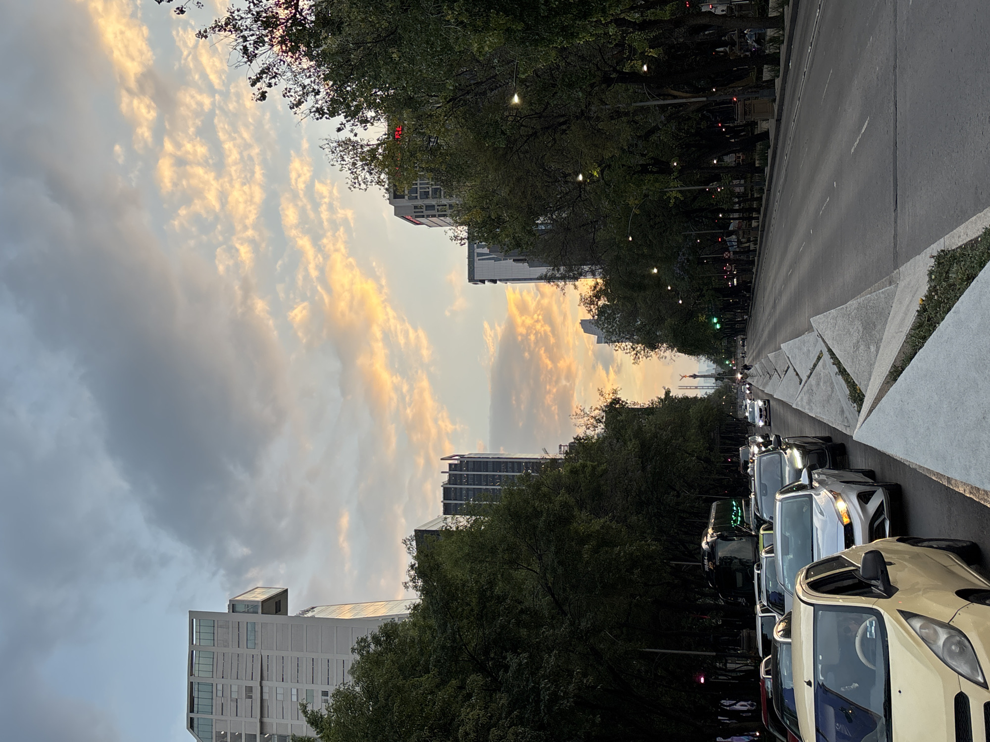 The image depicts a view along a wide avenue in Mexico City, Mexico, with buildings visible in the background and trees lining the sides of the street. The sky is filled with clouds, some with a golden hue, suggesting it might be either sunset or sunrise. Traffic is congested, with several cars lined up in the lane closest to the viewer. Streetlights and traffic signals are visible along the avenue. The architecture of the buildings suggests a modern urban landscape.