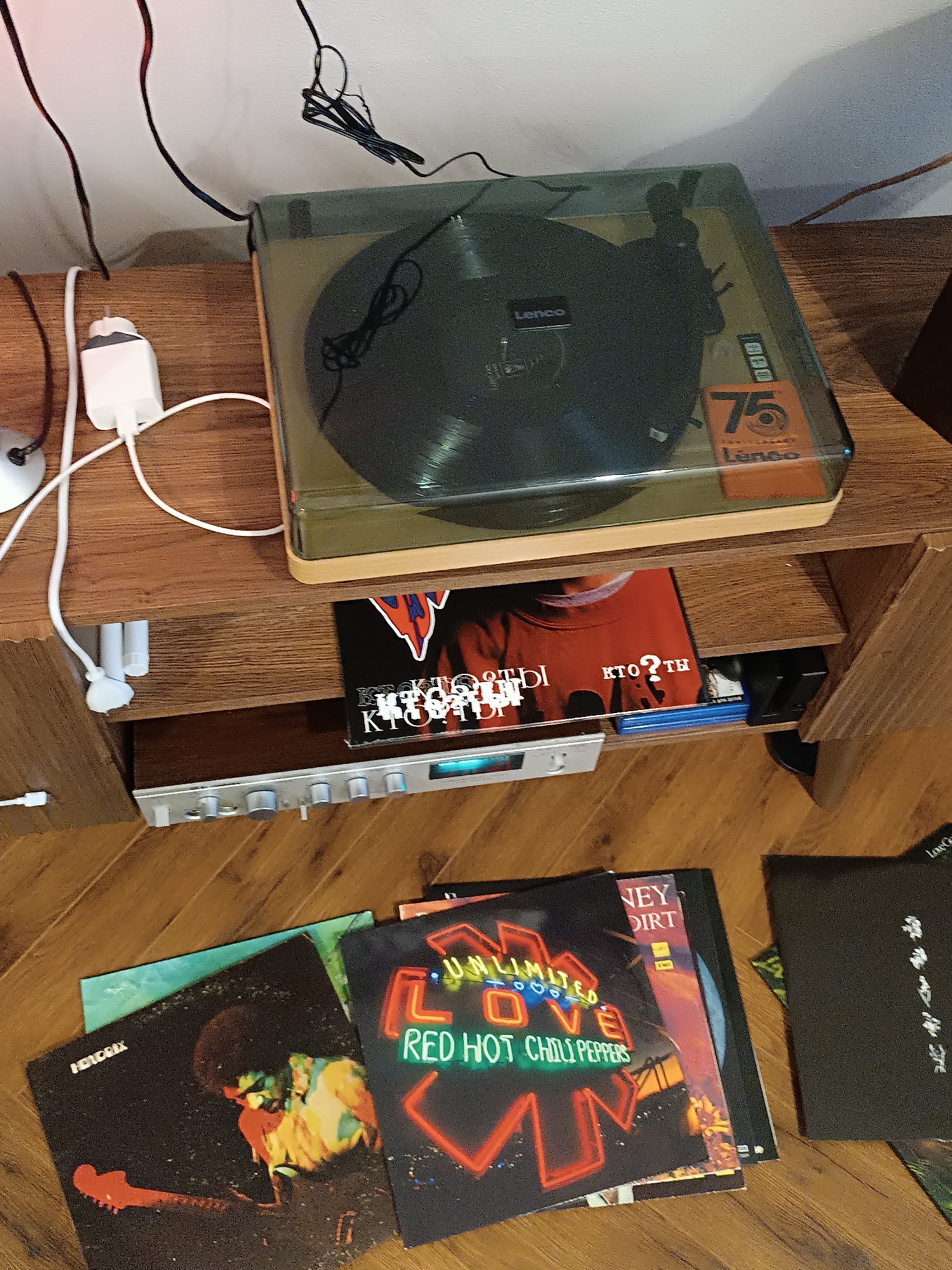 The image shows a setup of audio equipment on a wooden cabinet. On top of the cabinet is a Lenco turntable with a clear plastic lid. Below the turntable is a shelf that holds a Blu-ray disc and an audio component with silver knobs. On the floor in front of the cabinet, there are several vinyl records including albums by Red Hot Chili Peppers ("Unlimited Love") and Jimi Hendrix ("Hendrix"). The location is indoors, likely in a home. There are no people visible in the image.
