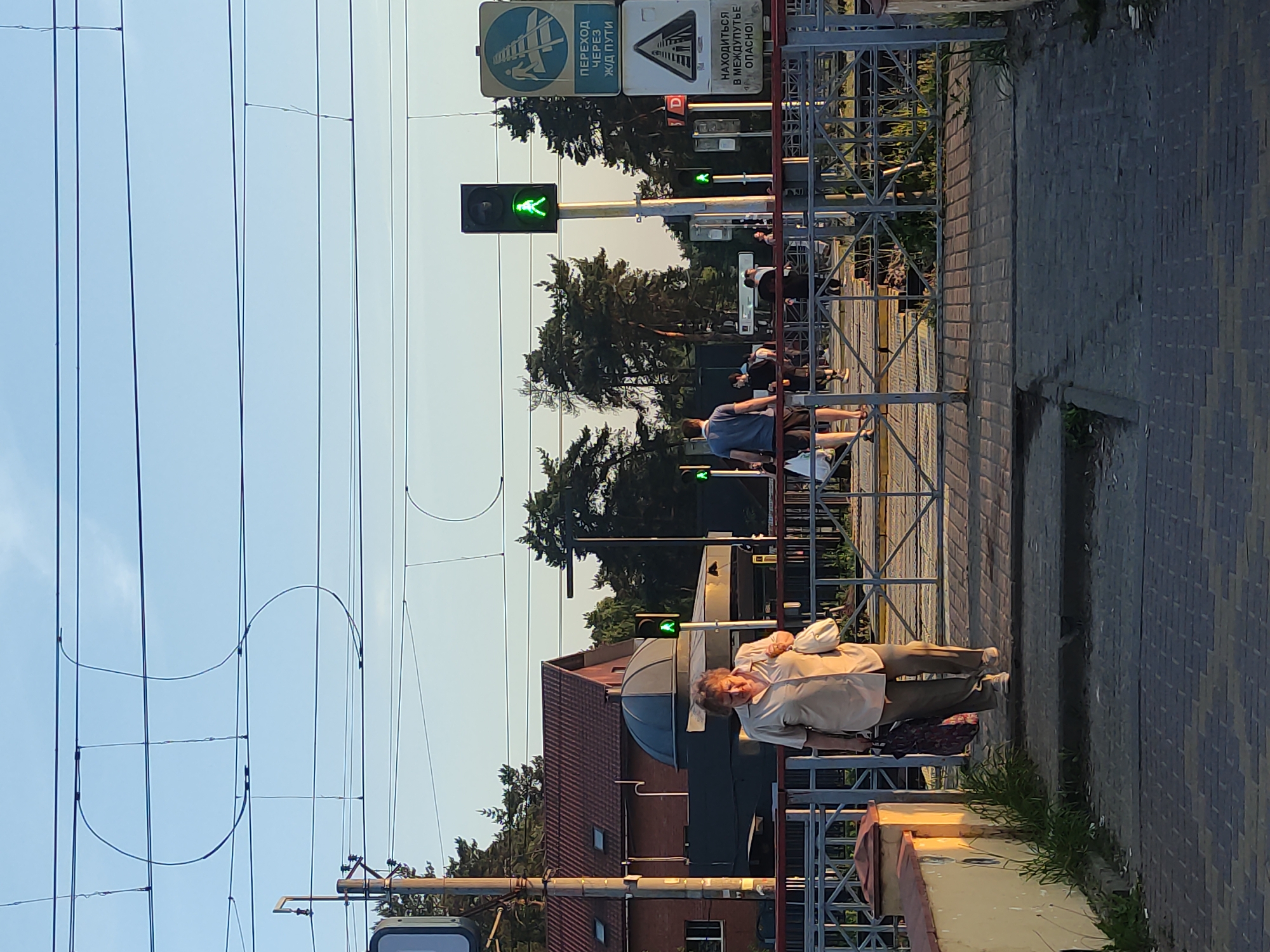 This image captures a pedestrian railway crossing in Kratovo, Russia, under a clear, bright sky. The scene is dominated by a multi-track railway line traversed by a paved pedestrian path with metal safety barriers and gates.
In the immediate foreground, a textured, dark grey brick-paved path transitions to worn concrete steps. To the left, a low, light-colored wall with a red cap is visible, bordered by grass.
An elderly woman, central in the frame, walks towards the crossing. She wears a light-colored, short-sleeved shirt and matching trousers, carrying a white handbag and a patterned dark bag. Ahead of her, a man in a dark blue t-shirt and shorts walks towards the tracks, carrying a white bag. Several other pedestrians are visible further along the crossing.
A prominent black pedestrian signal pole stands centrally on the crossing, displaying a green "walking man" symbol. Another green signal is visible further left, and one further right, indicating the crossing is open. To the right of the main signal, two informational signs in Russian are displayed: a blue rectangular sign indicating "CROSSING OVER RAILWAY TRACKS" (ПЕРЕХОД ЧЕРЕЗ Ж/Д ПУТИ) and a white triangular warning sign stating "BEING BETWEEN TRACKS IS DANGEROUS!" (НАХОДИТЬСЯ В МЕЖДУПУТЬЕ ОПАСНО!). A small red "D" sign is partially visible below the white sign.
In the mid-ground background, multiple railway tracks are visible beneath the pedestrian pathway. To the left, a reddish-brown brick building with a tiled roof and skylights stands behind a distinctive dark-domed structure. A wooden utility pole with electrical equipment is positioned in front of the brick building.
The background is filled with a dense line of green trees, likely conifers, suggesting a wooded area. Numerous dark overhead power lines, including characteristic S-shaped catenary wires, crisscross the clear, light blue sky.