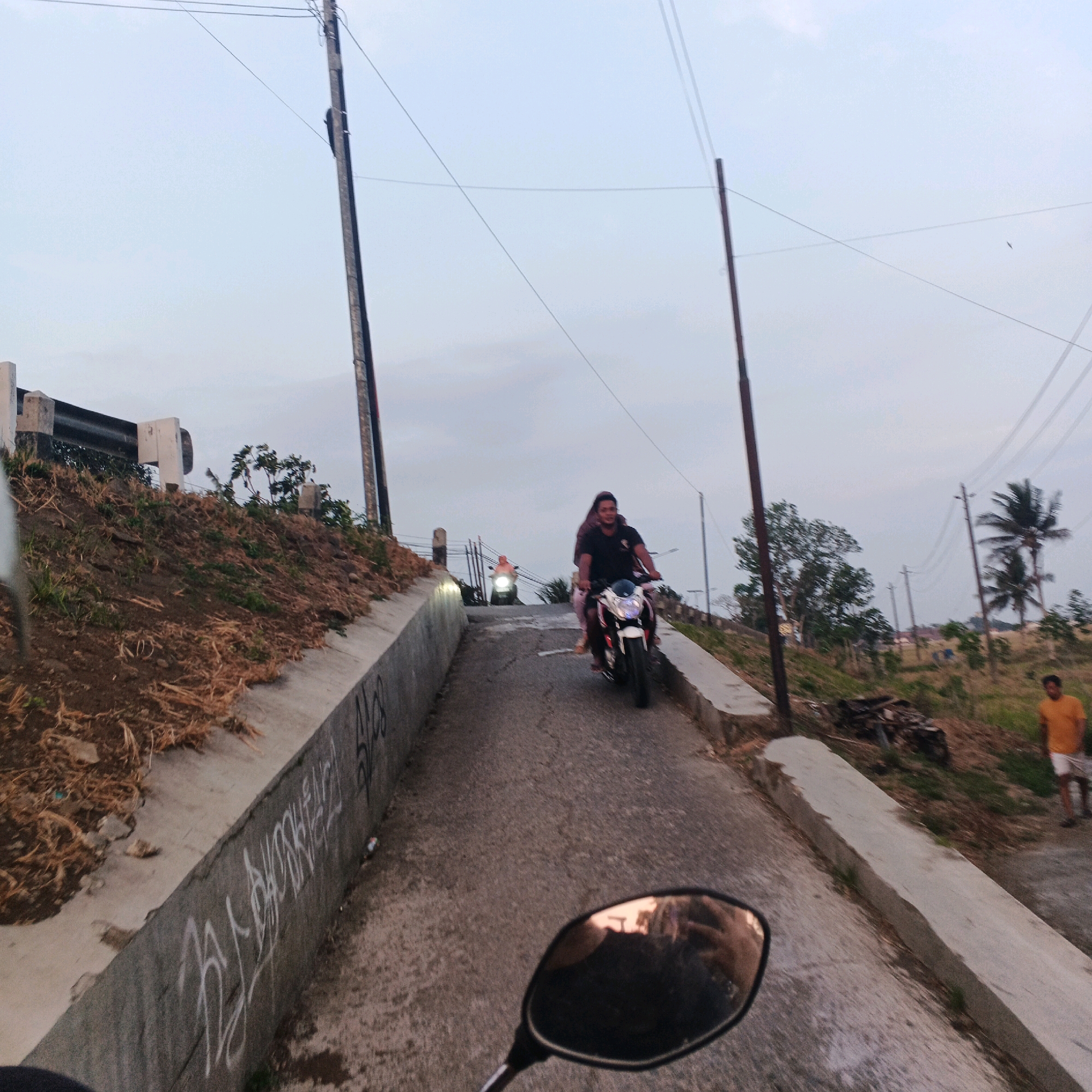 The image shows a slightly upward-sloping paved road in what appears to be a rural or semi-rural area of Kalikudi, Indonesia. Two motorcycles are visible on the road. The closest motorcycle, from which the photo is taken, has a rider and a passenger. The rider is wearing a black t-shirt and light-colored pants, and the passenger is wearing a dark-colored head covering and a light-colored garment. The second motorcycle is further up the road, with a rider wearing an orange shirt.
To the left of the road is a sloped embankment covered with dry grass and soil, bordered by a concrete retaining wall with graffiti. A metal guardrail is present at the top of the embankment. To the right of the road, there is a grassy area with scattered debris and several utility poles with overhead wires. In the background, there are trees, including palm trees, and a few buildings. The sky is overcast, suggesting it might be late afternoon or early morning. A person wearing an orange t-shirt and white shorts is visible in the distance on the right side of the frame.