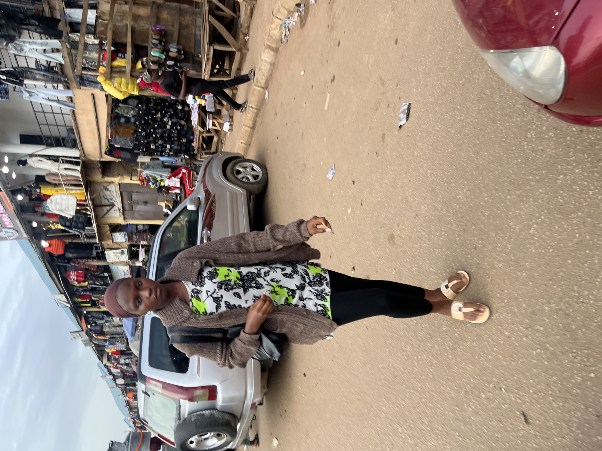 In the city of Bukuru, Nigeria, a young woman walks forward on a dusty, paved road. She is wearing a brown knitted cardigan over a white shirt with a green and black floral print, black leggings, and light-colored sandals. A black bag is slung over her left shoulder. In the background, stalls of clothing are visible, with items displayed on racks and shelves. A silver SUV is parked on the left, and a red car is partially visible in the foreground on the right. The sky is overcast, suggesting a cloudy day. The road appears to be part of a marketplace.