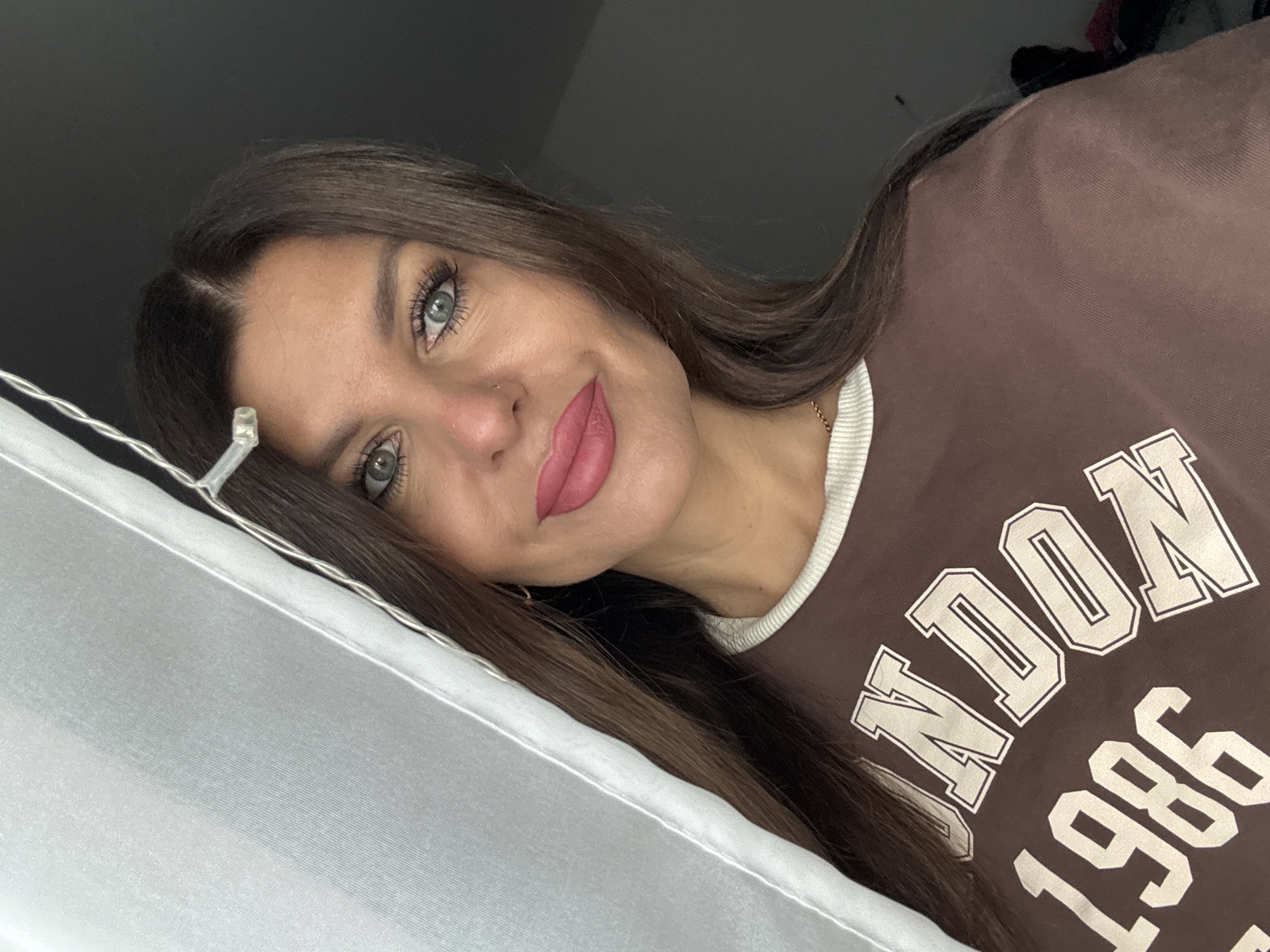 A woman with long, dark brown hair and blue-green eyes is looking at the camera. She is wearing a brown t-shirt with "LONDON 1986" printed on it in off-white lettering. Her lips are painted a matte pink. She has a subtle nose ring and a delicate gold chain around her neck. A sheer white curtain is draped to her left, obscuring a portion of the background. To the right of the woman, the background is dark and appears to be an indoor space. No specific location in Chaikovsky, Russia can be identified.