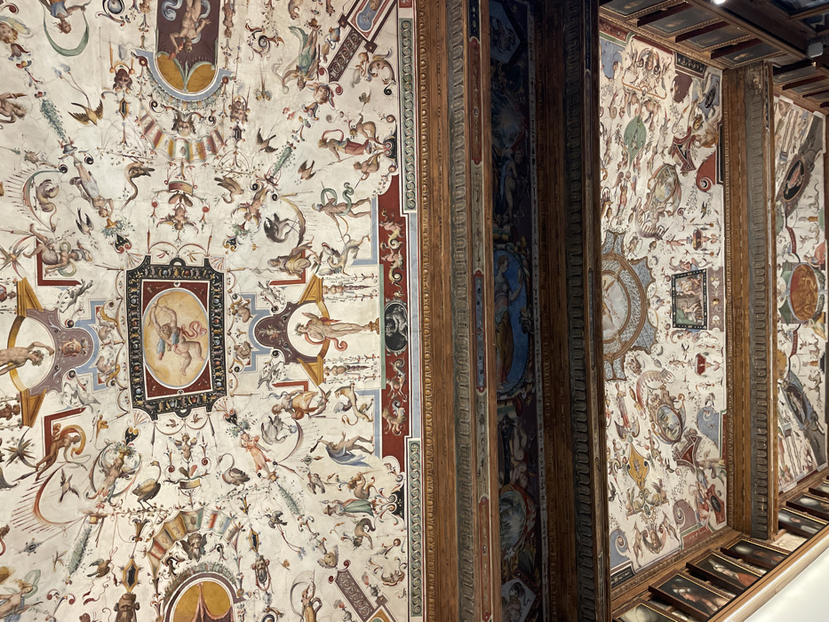 The image displays an upward view of an elaborately decorated ceiling within the Palazzo Vecchio in Florence, Italy. The primary visual is a vast frescoed ceiling, rendered in the grotesque style, featuring a dense arrangement of figures and motifs on a light background. Prominent elements include numerous winged putti, mythological figures, various human forms (some nude, some draped), fantastical beasts, birds, and animals, intertwined with intricate floral patterns and architectural ornamentations.
Specific details visible on the ceiling include an oval framed panel in the upper section depicting a winged, fair-skinned figure, possibly Cupid, drawing a bow. Below this, a rectangular panel features a standing, semi-nude male figure within an ornate niche, likely a deity. The fresco's color palette comprises muted tones of red, blue, green, yellow, and brown, contributing to its aged aesthetic.
Below the main frescoed ceiling, a series of heavy, dark brown wooden beams with intricate carvings and subtle painted details (including red, blue, and gold accents) traverse horizontally. These beams define a coffered structure, with recessed panels between them also containing painted decorations. Further below, a portion of a lower, similarly decorated ceiling is visible, continuing the grotesque motifs. On the far left and right edges of the frame, sections of walls are discernible, adorned with multiple framed portrait-style paintings arranged vertically, suggesting a richly appointed interior space. The overall scene depicts a complex, multi-layered decorative scheme from the High Renaissance or Mannerist period.