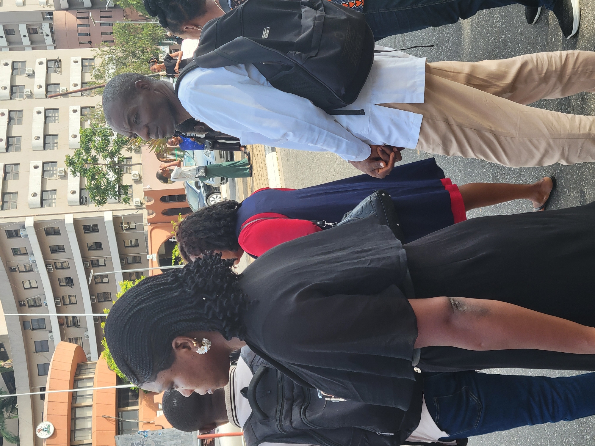 Several people are standing outside on a paved area in Abuja, Nigeria. The individuals appear to be attending an event or gathering. A man wearing a white shirt, khaki pants, and a black backpack stands with his hands clasped. A woman in a black top with braided hair and flower-shaped earrings is also in the foreground. Another woman is wearing a red top and a dark blue skirt. Buildings are visible in the background, including a multi-story building with rows of windows and air conditioning units. A tree and a car are also visible.