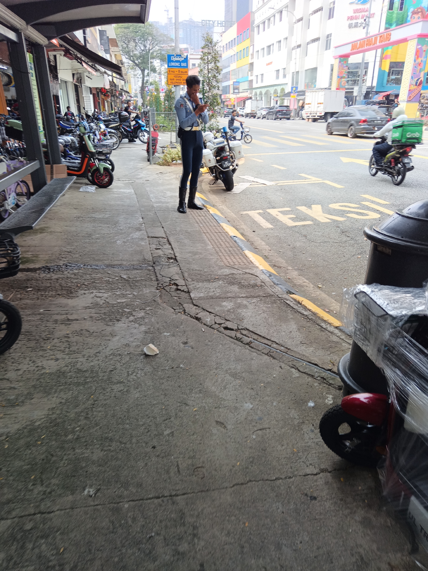 The image depicts a street scene in Kuala Lumpur, Malaysia, specifically near the area of Jalan Haji Taib and Lorong Bas. In the foreground, a person in a blue uniform with black boots and a gold belt is standing on a cracked concrete sidewalk, looking at their phone. To the right, a black trash bin is partially visible, wrapped in plastic. Several electric scooters and motorbikes are parked along the sidewalk. In the background, traffic is visible on a street marked with yellow "TAKSI" (taxi) symbols. Buildings of various colors line the street, with some featuring signage in Malay and Chinese. A Grab food delivery rider on a motorbike is also visible in the traffic. The environment appears to be a busy commercial area with a mix of pedestrian and vehicle traffic.