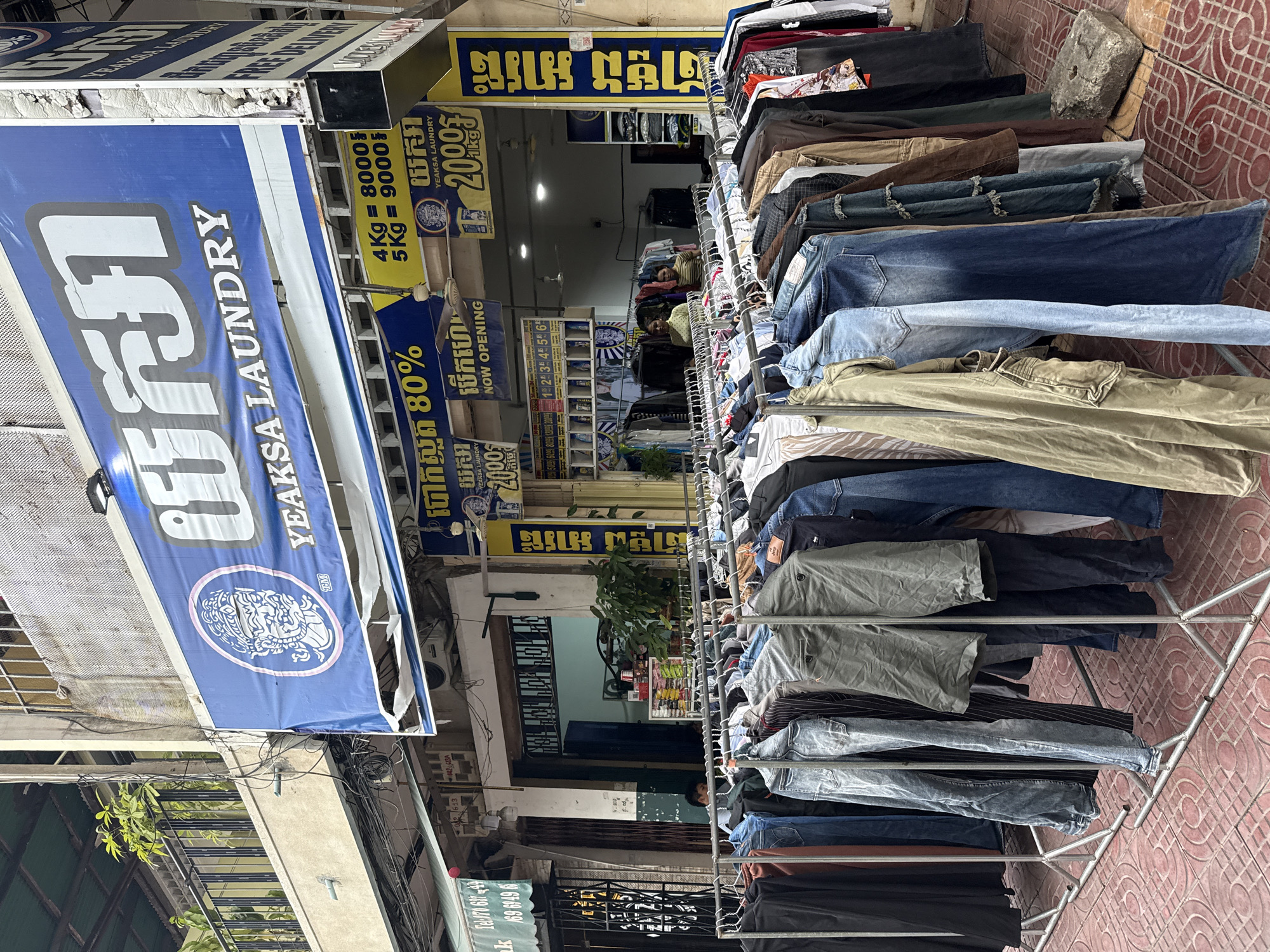 This is a street-level, medium-shot photograph taken in front of a laundry business in Khan Daun Penh, Cambodia. The facade of the business is dominated by a large blue sign with white text that reads "YEAKSA LAUNDRY" in English, along with Khmer script. Below this main sign, there are yellow signs advertising prices and services, including "80%" and "20,000 / 1KG." A rack of assorted clothing, including jeans, jackets, and pants, is displayed on hangers outside the establishment, extending across the foreground of the image. The clothing is arranged on a metal garment rack, and the ground is paved with red tiles in a circular pattern. In the background, the entrance to the laundry business is visible, with two women standing inside. One woman appears to be wearing a yellow top, and the other a striped shirt. A ceiling fan is visible above them. The overall environment suggests a commercial street in a Southeast Asian city.