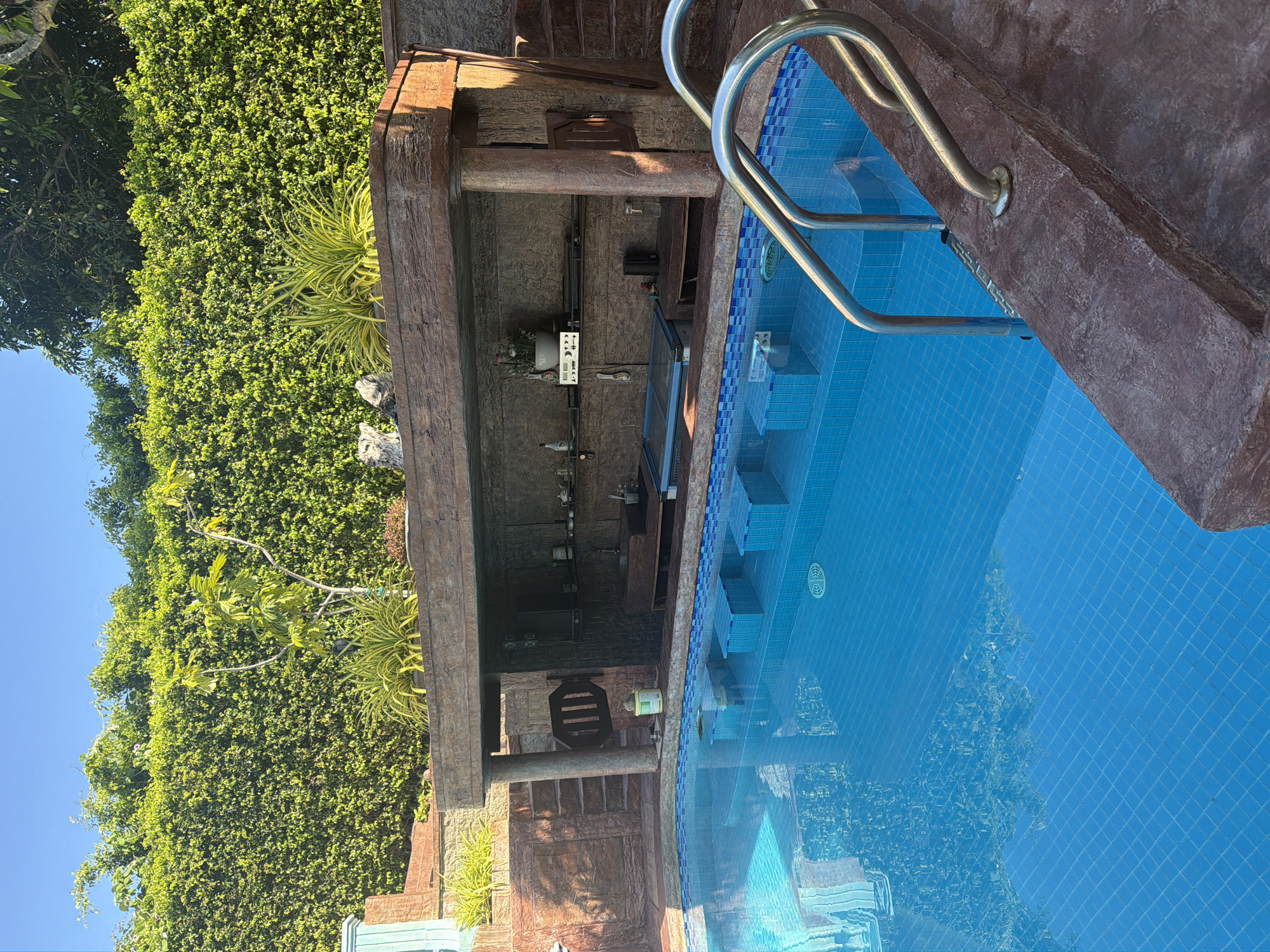 The image depicts a swimming pool with a built-in bar area. The pool is tiled with blue squares and has a stainless steel ladder for entry and exit. Adjacent to the pool is a bar structure with wooden pillars and a counter stocked with bottles and equipment. A dense green wall of foliage forms the backdrop of the bar. The image was taken in Bac Nha Trang Ward, Vietnam.