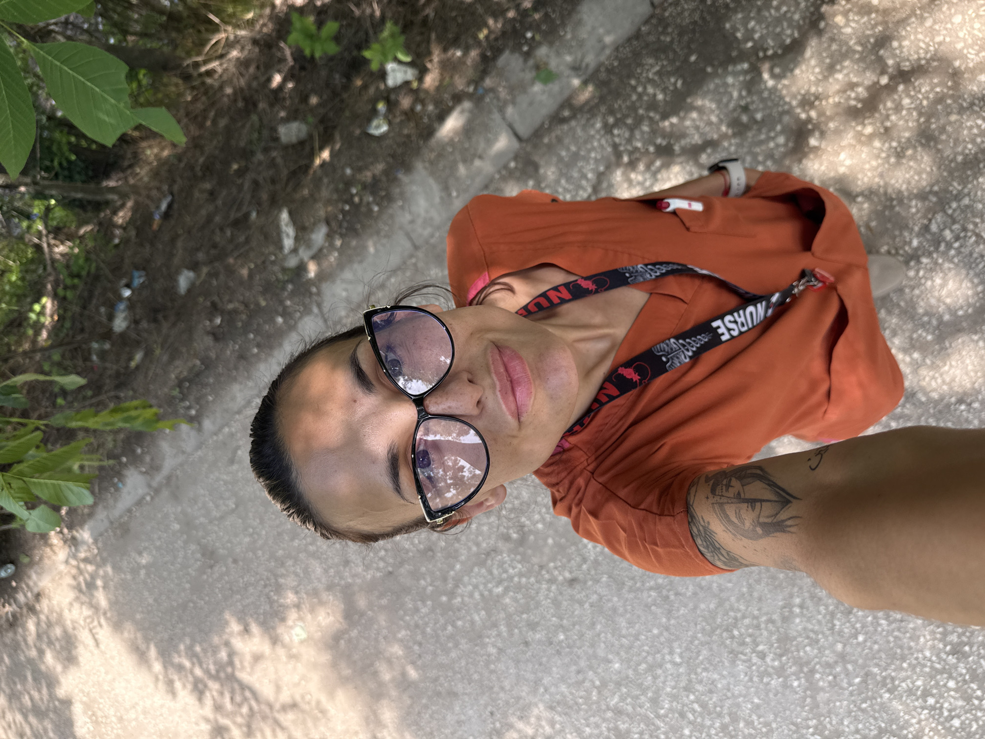 An adult female is captured in a high-angle selfie, taken outdoors in Pleven, Bulgaria. She has dark hair pulled back and wears black-framed eyeglasses with reflective, slightly purple-tinted lenses. Her face shows freckles and a faint smile. She is dressed in an orange-red V-neck medical scrub top, with a black lanyard emblazoned with "NURSE" in white hanging around her neck. A pen with a red cap is visible in her left scrub pocket, and a white smartwatch is on her left wrist. A prominent black-ink tattoo depicting two stylized female faces joined at the center is visible on her left forearm, and a smaller dark, curvilinear tattoo is on her upper right arm. She holds the camera directly towards herself, looking upwards.
The environment shows a light-colored, possibly asphalt or gravel, path beneath her. To the right of the path, a dirt embankment covered with green foliage and littered with various debris, including plastic bottles and wrappers, is visible. Overhead, green leaves from a tree cast dappled shadows across the scene and the subject's face, indicating bright sunlight.