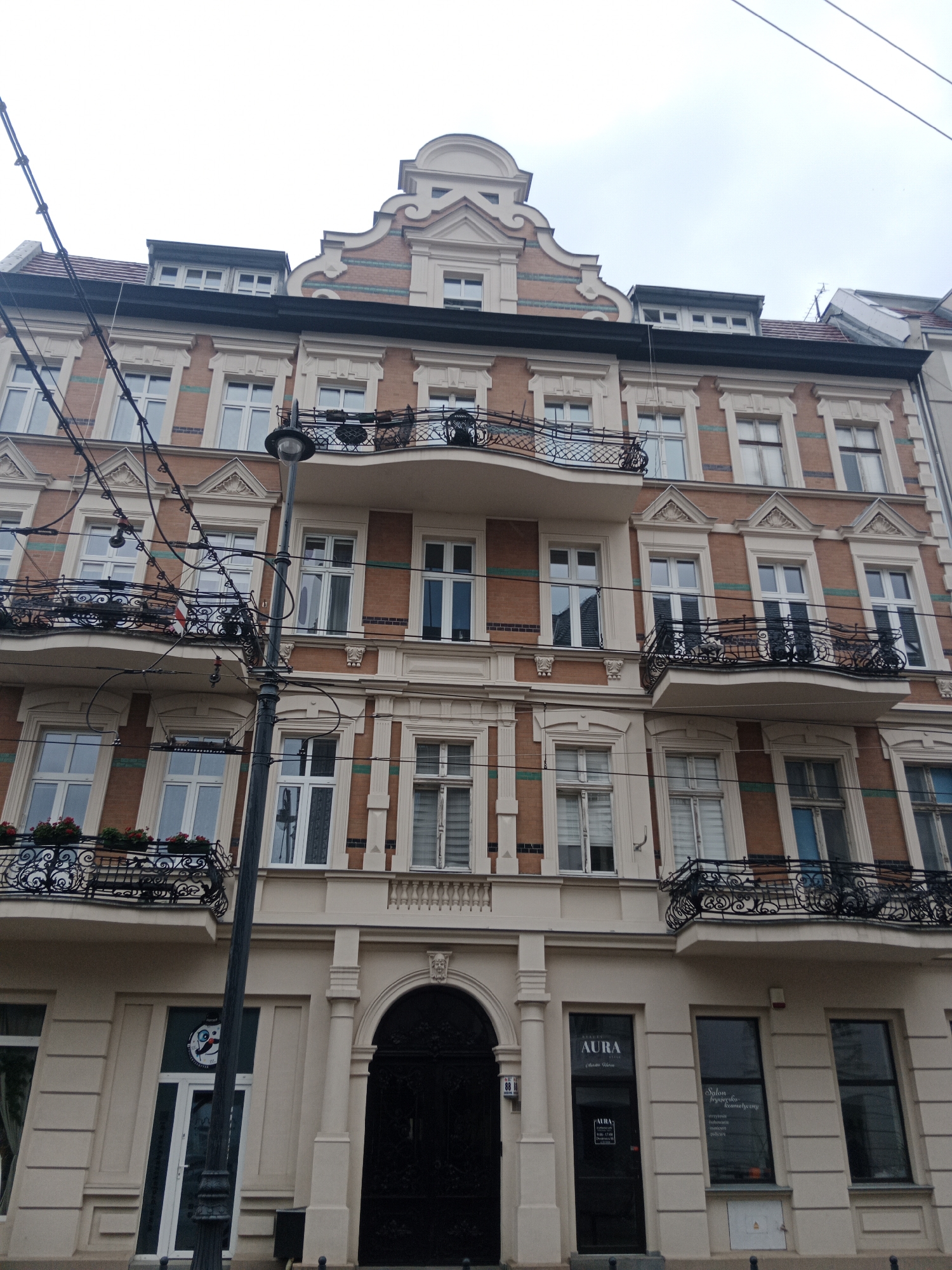 The image shows a building in Bydgoszcz, Poland, likely a residential or commercial property. The building has an ornate facade with red brick and cream-colored decorative elements. It features multiple stories with numerous windows, many of which are arched at the top. There are wrought-iron balconies with intricate designs on several levels. A prominent black lamppost stands in front of the building, along with electrical wires crisscrossing overhead. The ground level has an entrance with a large black door and a sign above it that reads "AURA." To the right of the entrance, there are shop windows displaying text that appears to be for a hair salon, with the words "Salon fryzjersko-kosmetyczny." The overall scene depicts an urban setting with a vintage architectural style.