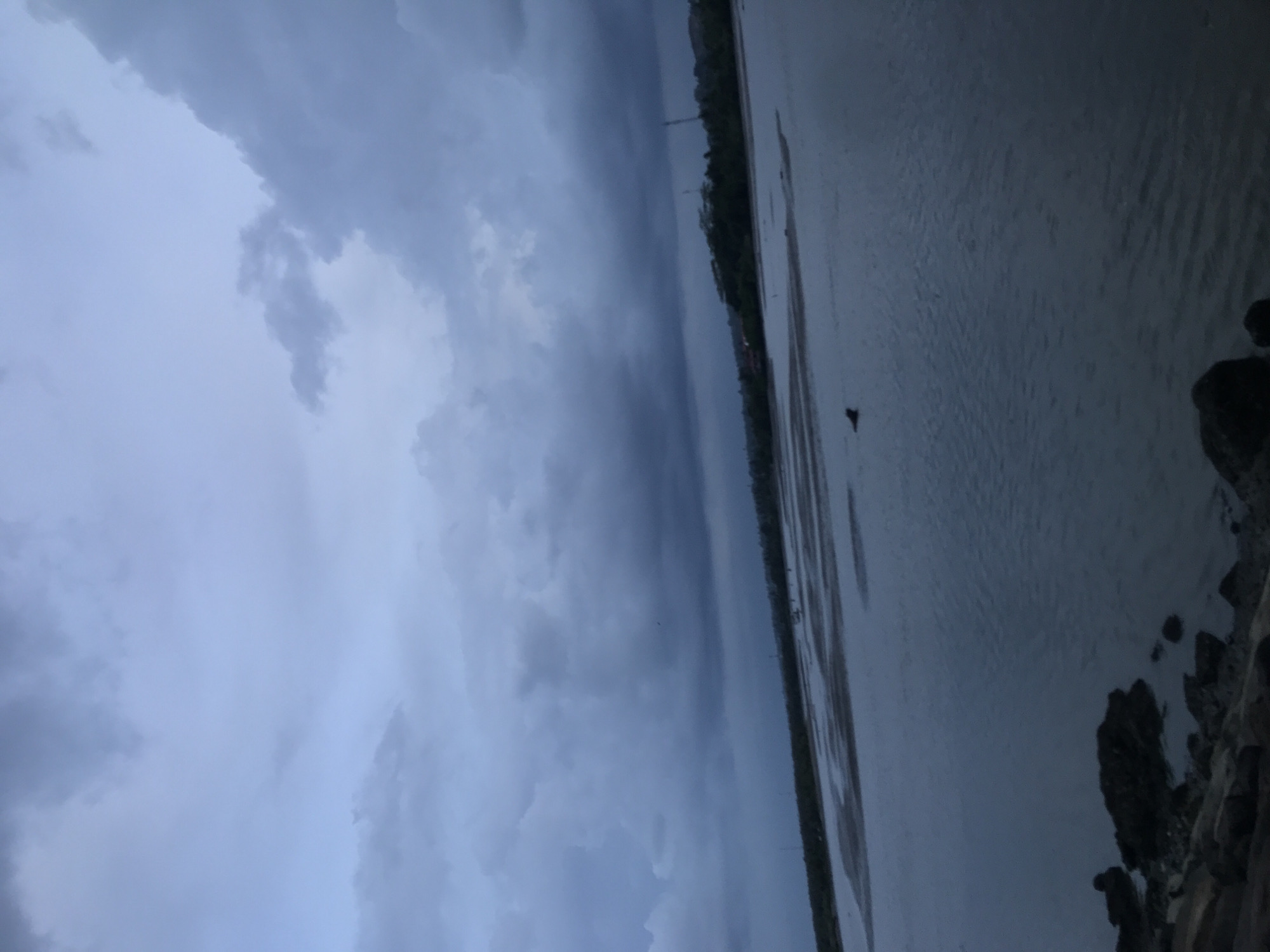 The image shows a body of water, likely a sea or a wide river, under a dark and cloudy sky. The water is gray, reflecting the somber atmosphere. A stretch of land, possibly a coastline or riverbank, runs along the horizon. Some rocks are visible on the bottom right of the frame. The overall scene suggests an overcast day, with the possibility of rain. The location is in the city of Lhokseumawe, Indonesia.