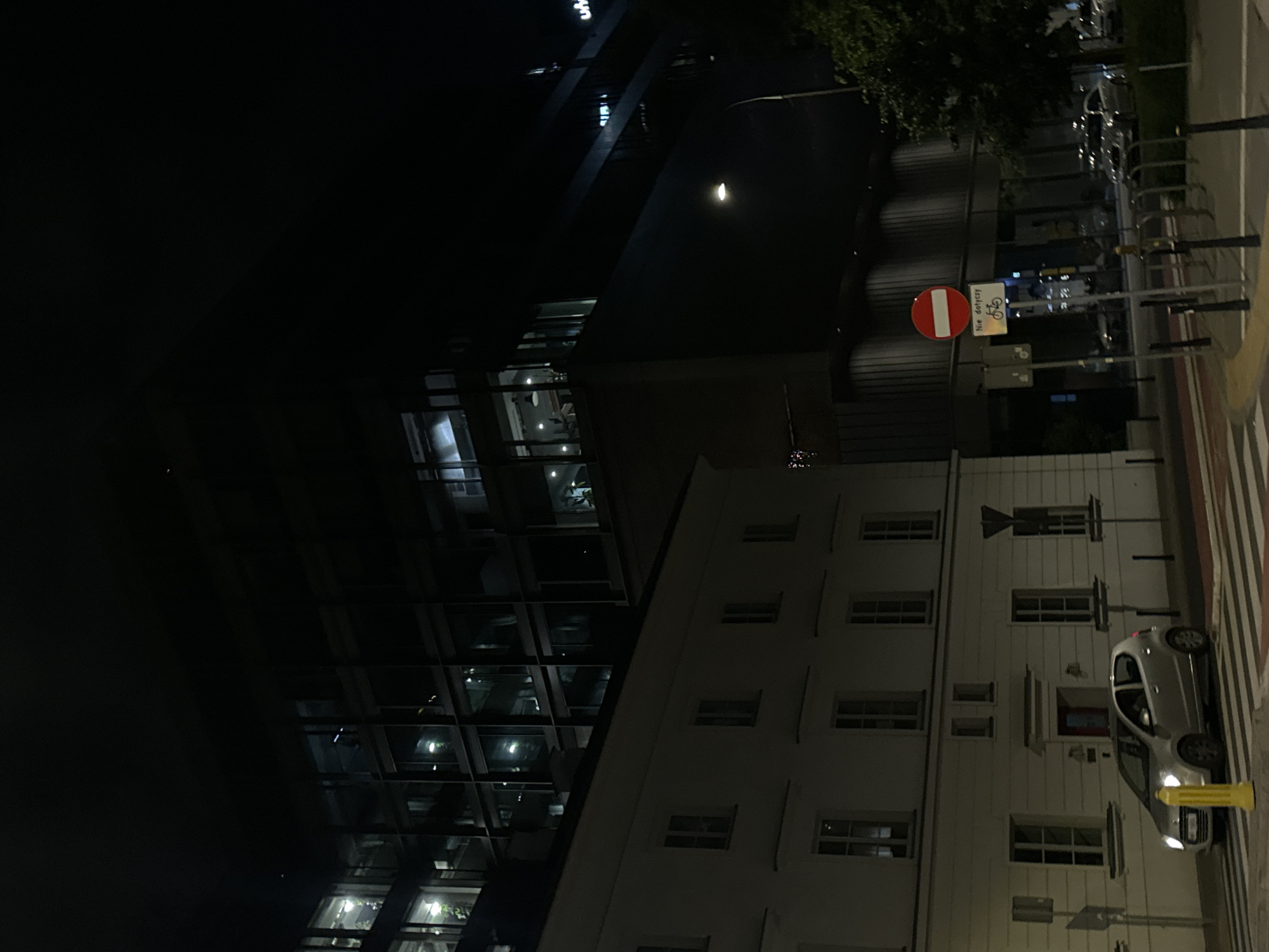 At night in an urban environment, a modern glass skyscraper intersects with a traditional white building. A no-entry traffic sign is visible on the side of the road with pedestrian crossing markings. A parked silver car is situated near a yellow fire hydrant.