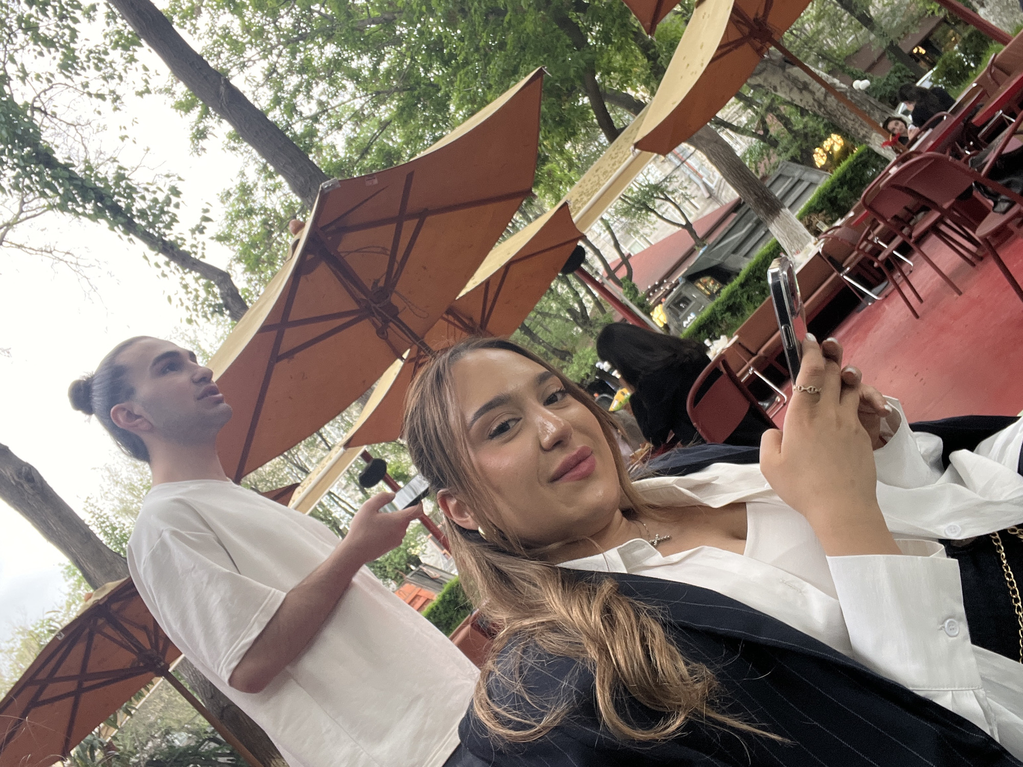 The image appears to be taken outdoors at a cafe or restaurant in Yerevan, Armenia. The weather seems pleasant as indicated by the outdoor setting. There are several orange parasols providing shade. A woman is sitting in the foreground, holding a smartphone, possibly taking a selfie. She is wearing a white collared shirt and a dark blazer with a gold necklace. A man is standing behind her to her left, wearing a white T-shirt and holding a phone as well. In the background, red chairs and tables are visible, along with other patrons of the establishment. Trees surround the area, providing a natural setting. The city's architecture can be glimpsed between the trees and parasols.