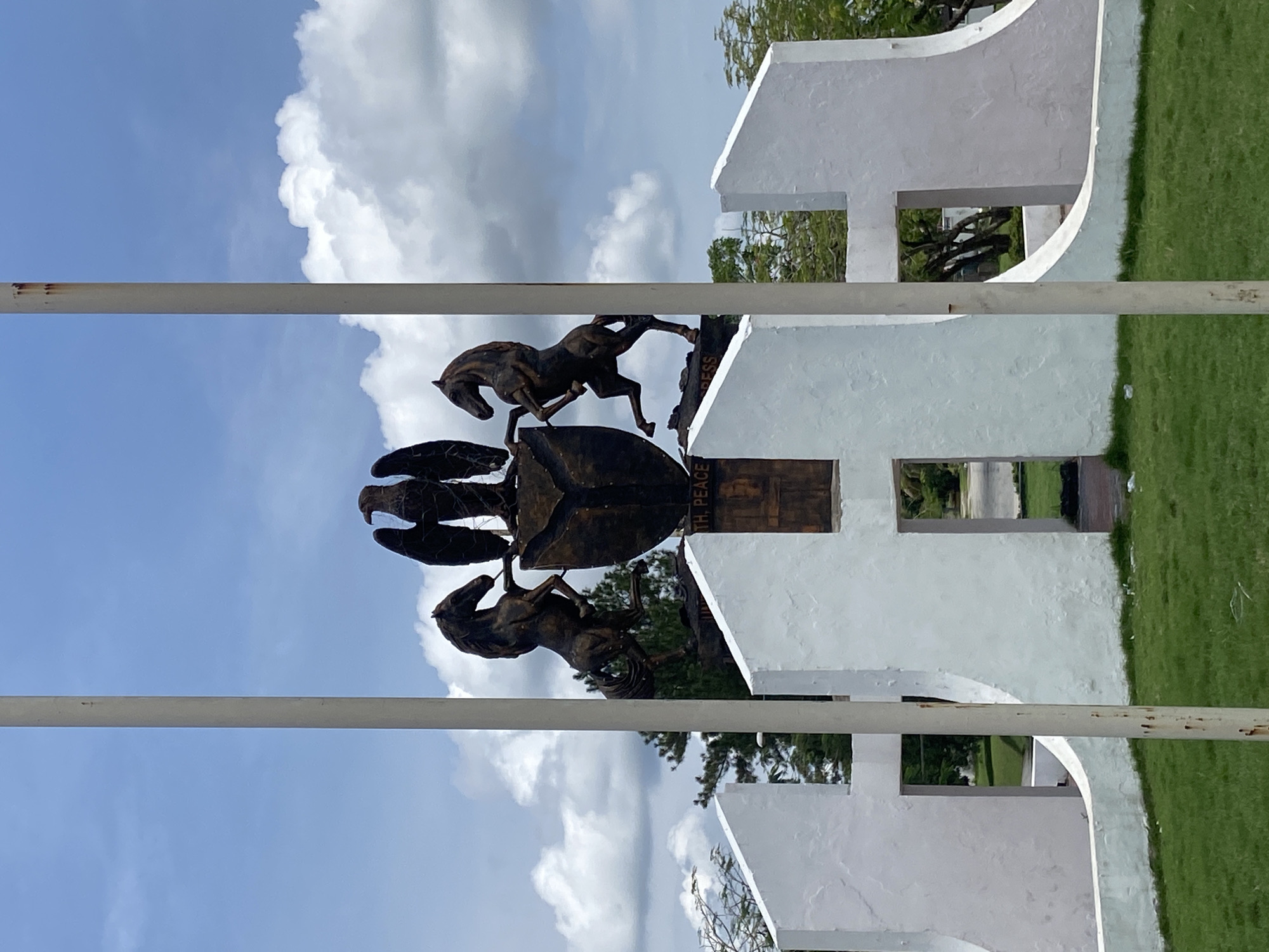 The image shows a monument in Calabar, Nigeria. It consists of a white, letter 'H' shaped structure with rectangular openings and a bronze coat of arms in the center. The coat of arms features two horses, one on each side, flanking a shield and a pair of winged hands on the shield. A metal bar is visible in the foreground. The background shows a blue sky with clouds and some green vegetation.